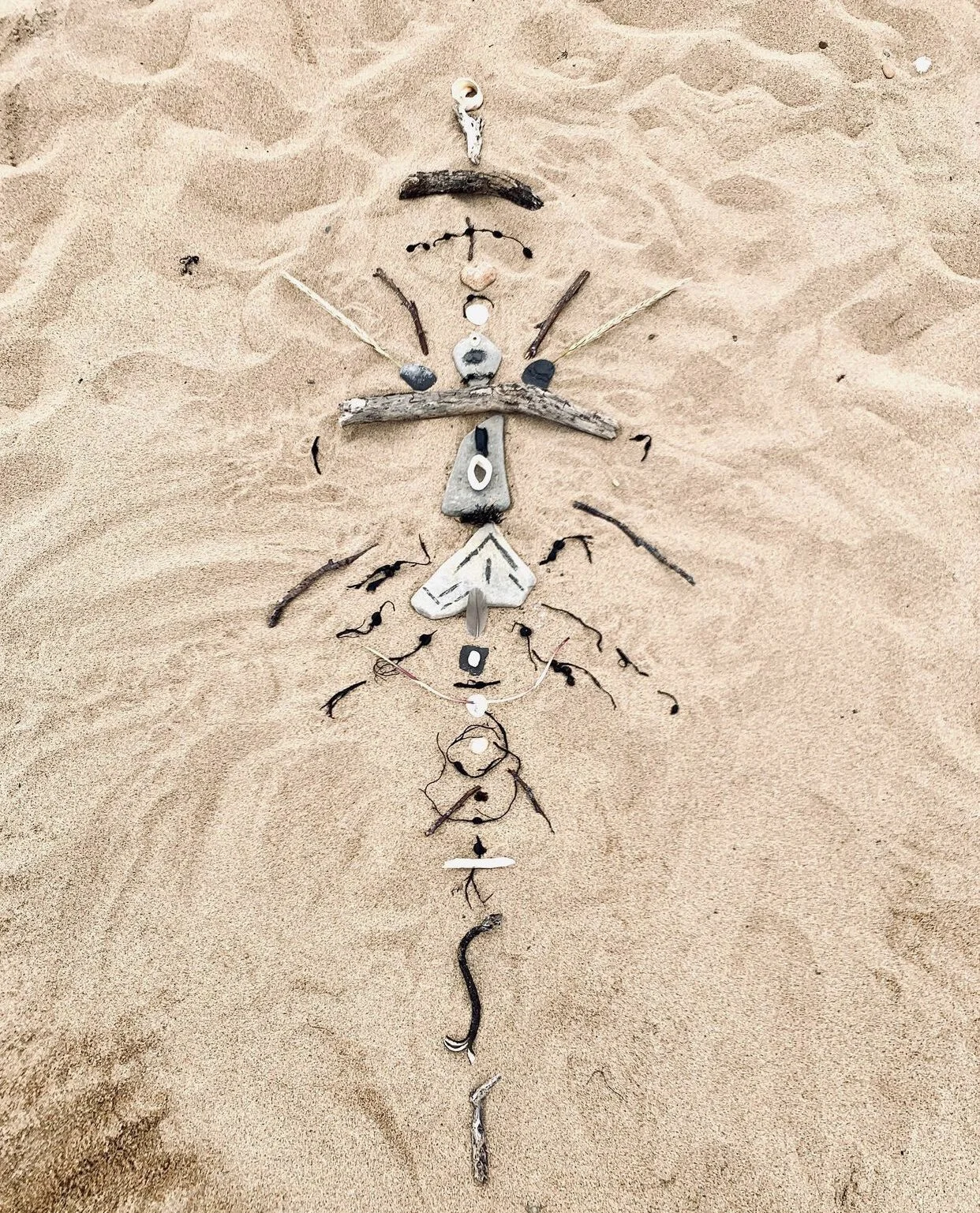 Arrangement of various small objects, including shells, stones, sticks, and shells, on sandy beach forming a human-like figure with outstretched arms, a head, and a body.