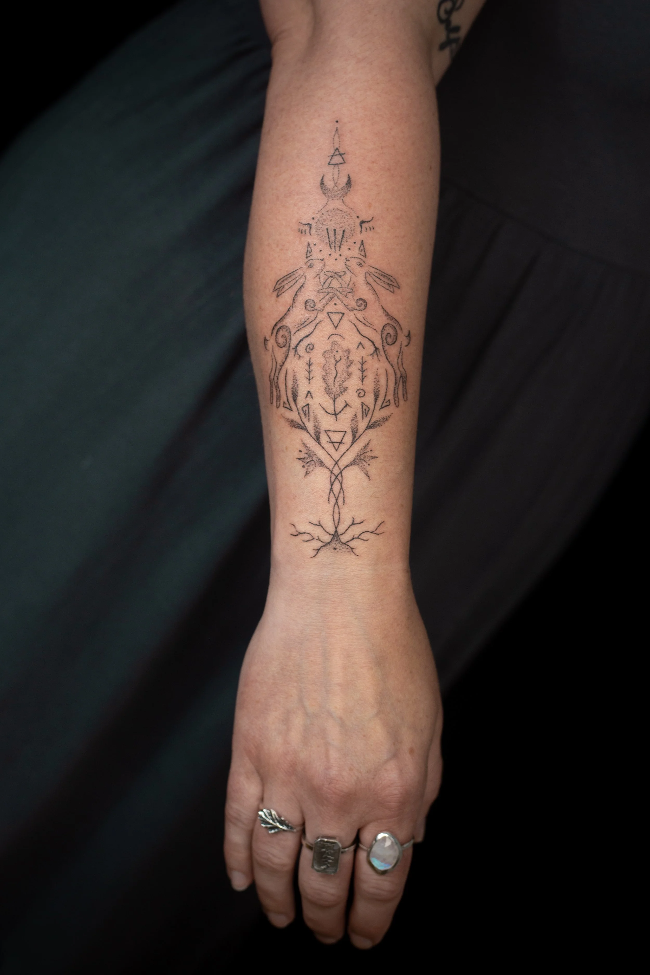 A person's forearm with a detailed black ink tattoo of two rabbits, geometric shapes, and nature-inspired elements.