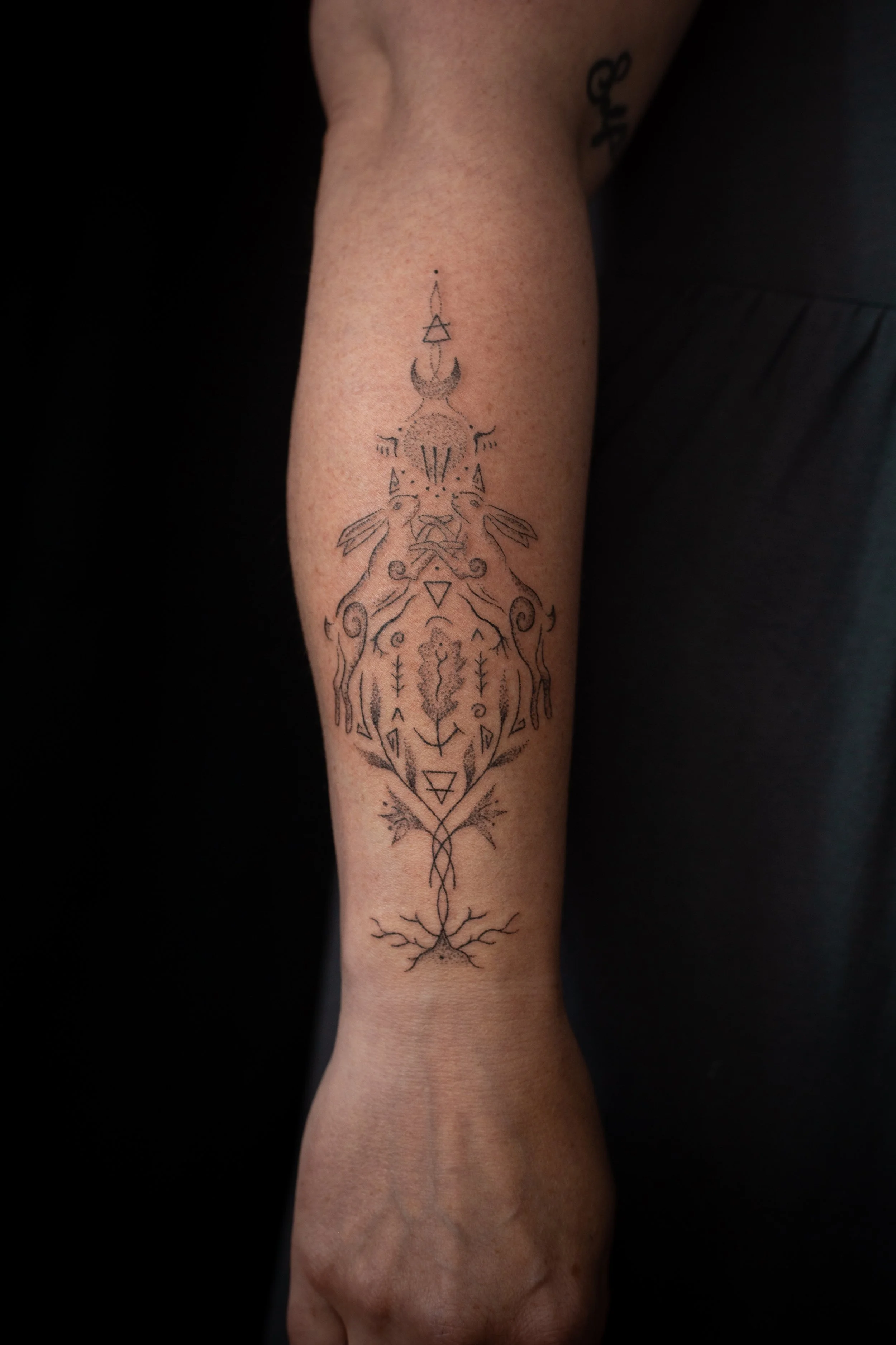A tattoo on a person's forearm featuring a symmetrical black ink geometric and animal design with a central tree-like element and two stylized animals facing each other.