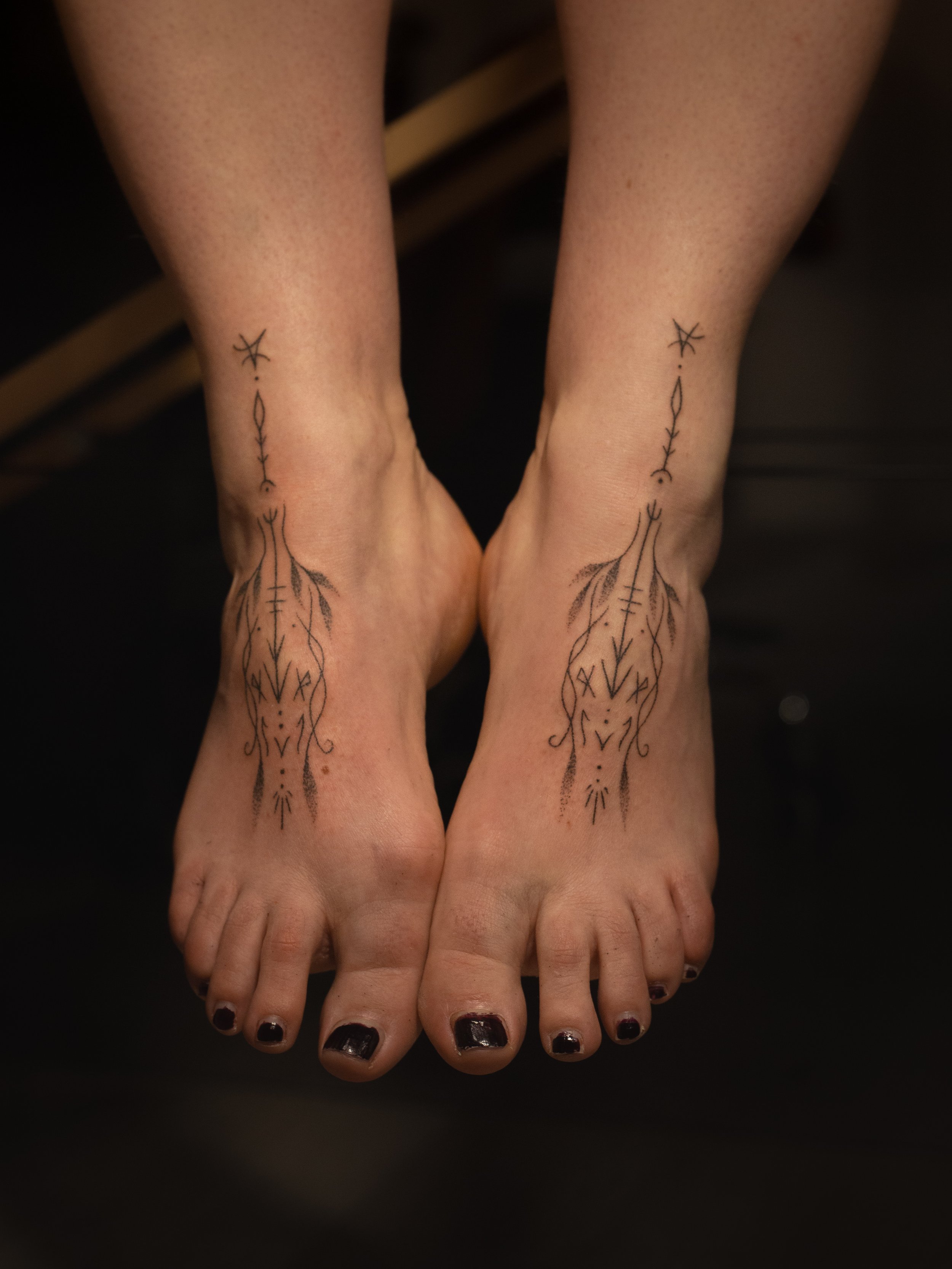 A person with black nail polish on their toenails, showing tattoos on both ankles, with intricate line designs resembling abstract or botanical patterns.