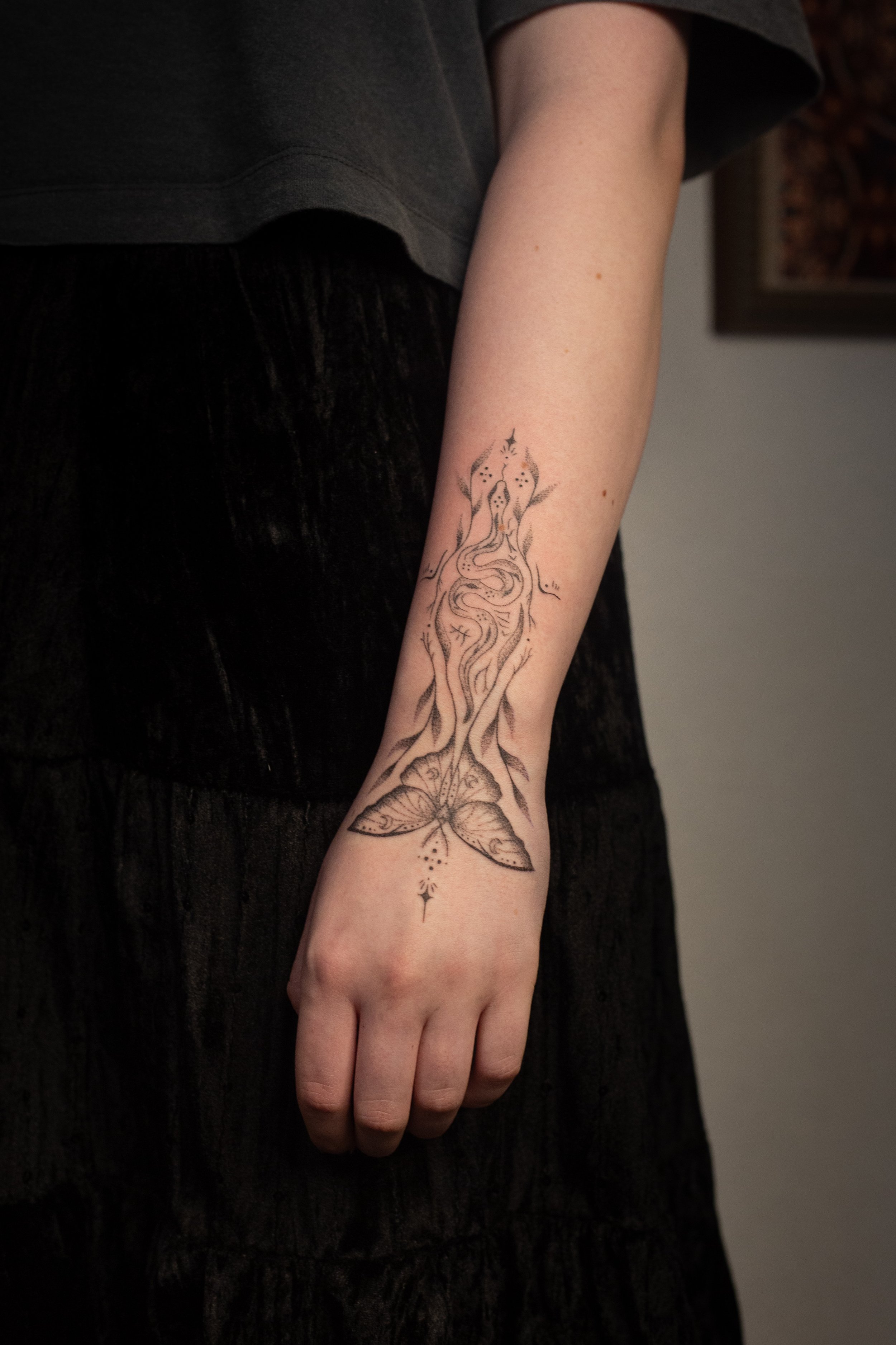 A person's arm with a detailed black tattoo of a bird, wind, and floral elements. The person is wearing a dark-colored short-sleeve shirt and black pants, with the background showing a wall with a framed picture.
