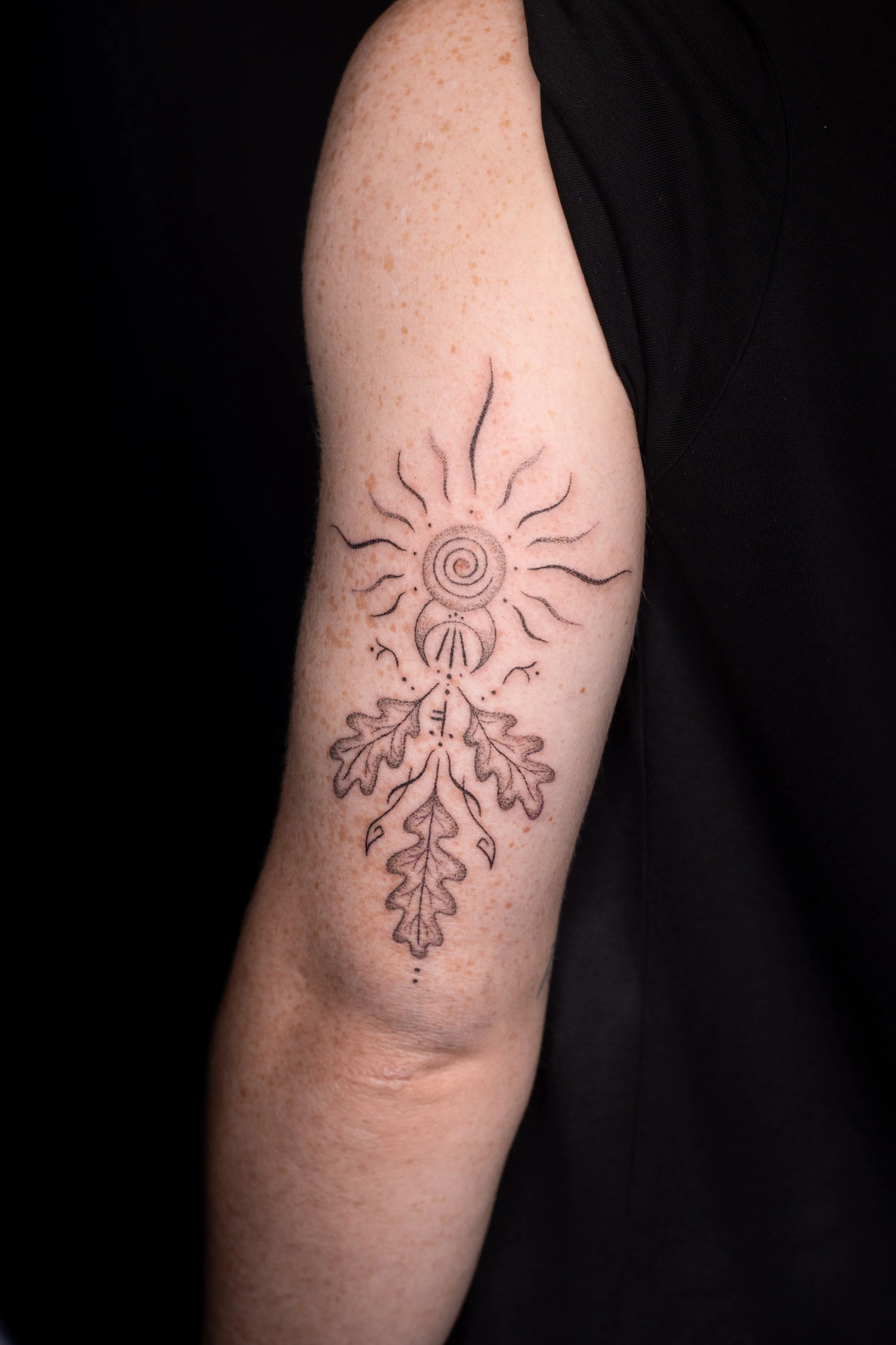 A person's arm with a sun and moon tattoo featuring a spiral sun with wavy rays and a crescent moon with small dots, surrounded by leaf and floral designs, against a black background.