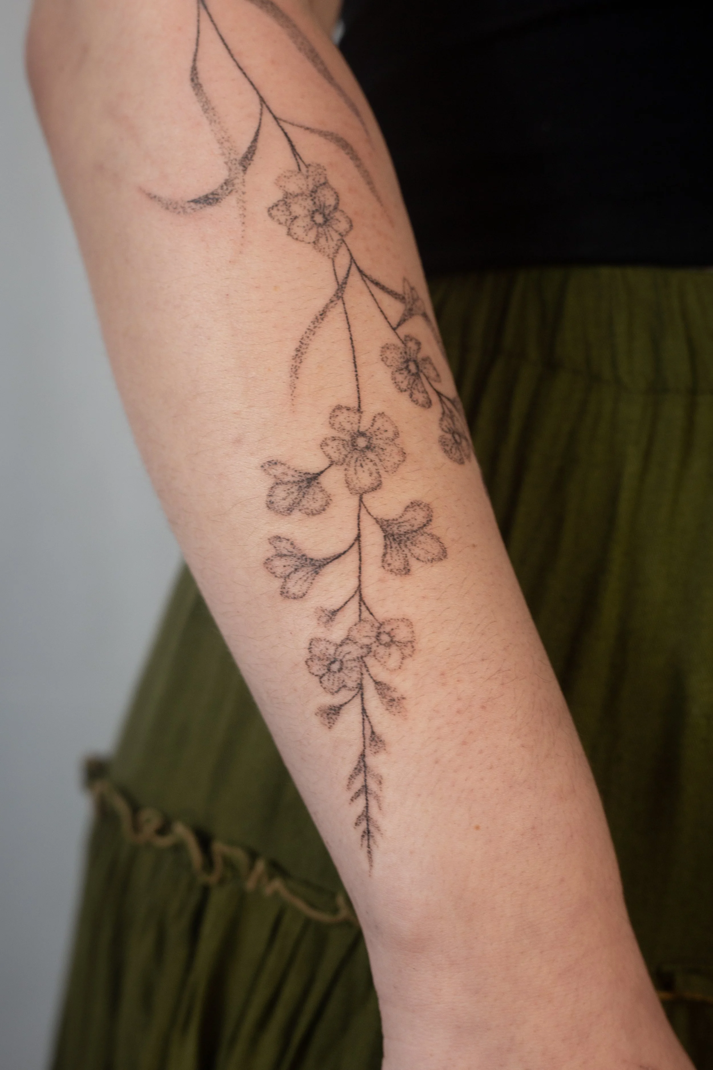 A black outline tattoo of a vertical floral vine with multiple flowers and leaves on a person's forearm.