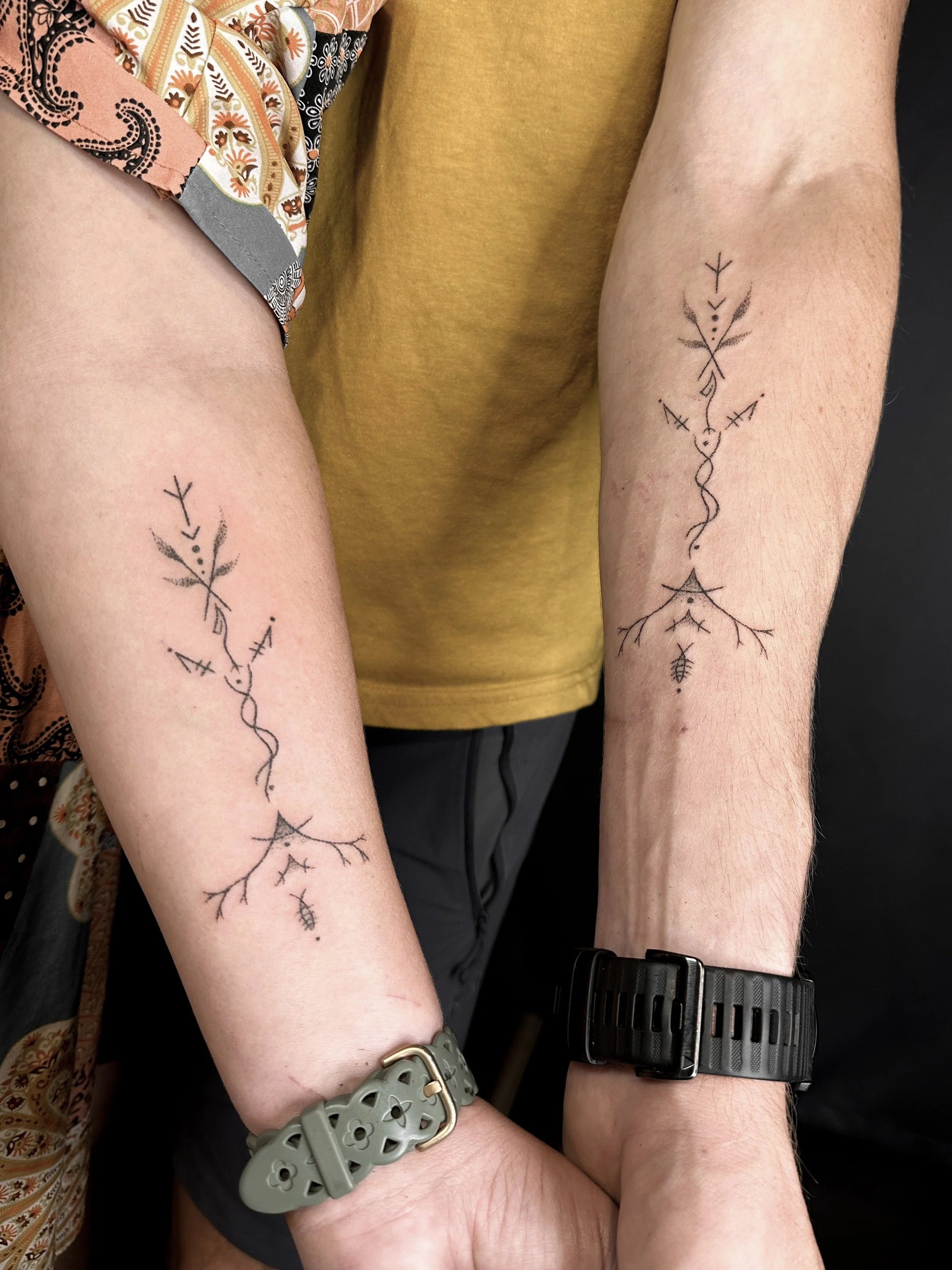 Two people with matching minimalist black line tattoos on their forearms, wearing watches and jewelry, holding hands.