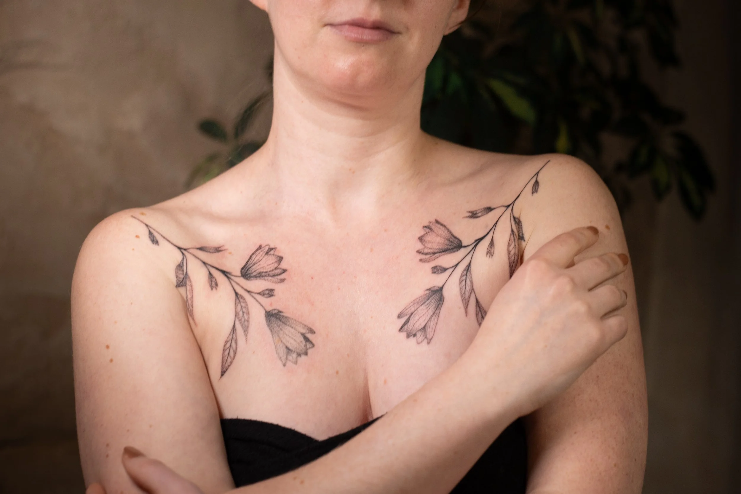 A close-up of a woman's upper body showing her bare shoulders and partial face. She has a floral tattoo on her chest and shoulder, and is wearing a black strapless top. Her arm is crossed over her chest with her hand resting on her other shoulder.