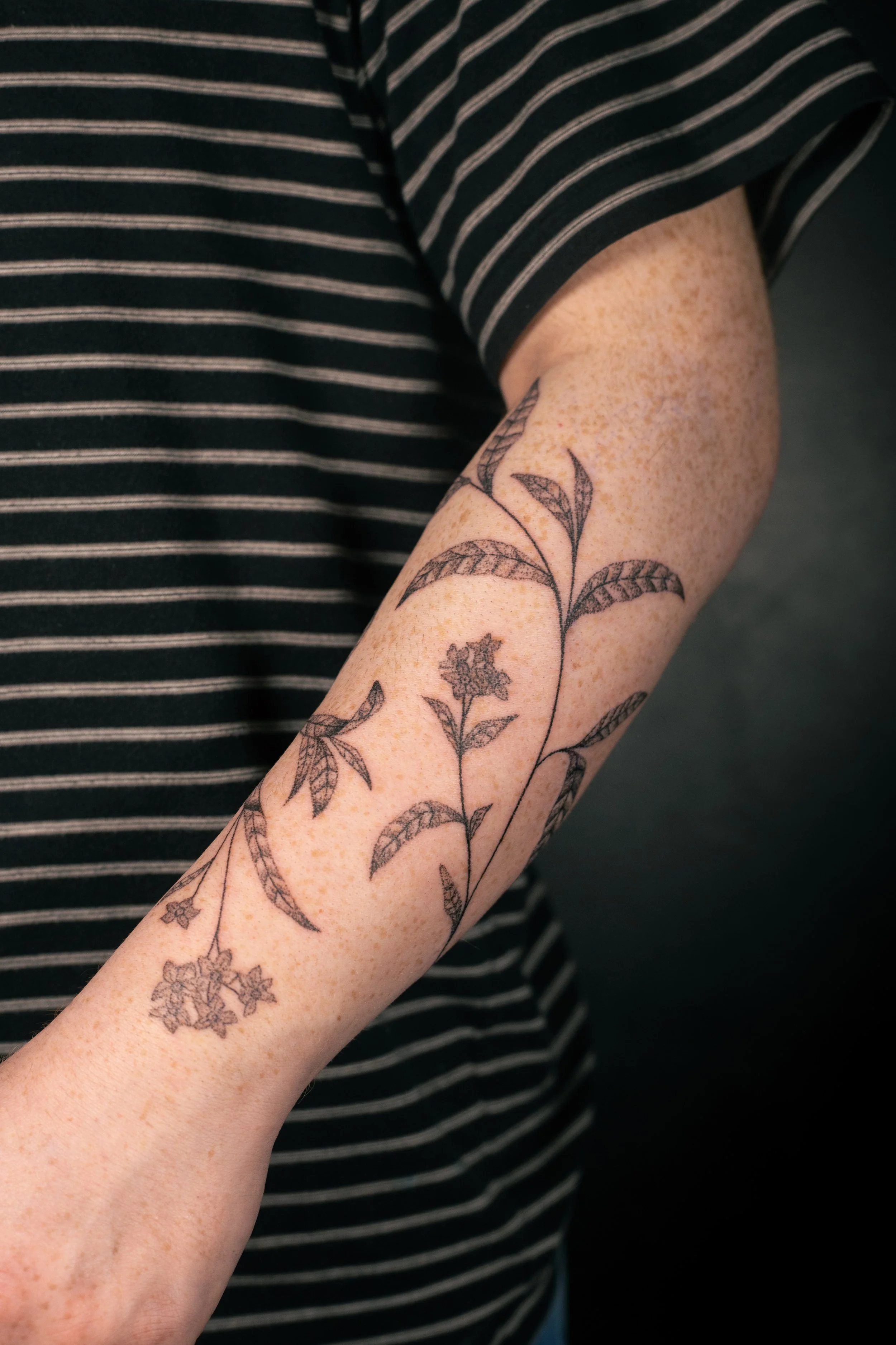 A person's arm with a floral tattoo featuring leaves and flowers, wearing a black and white striped short-sleeve shirt.
