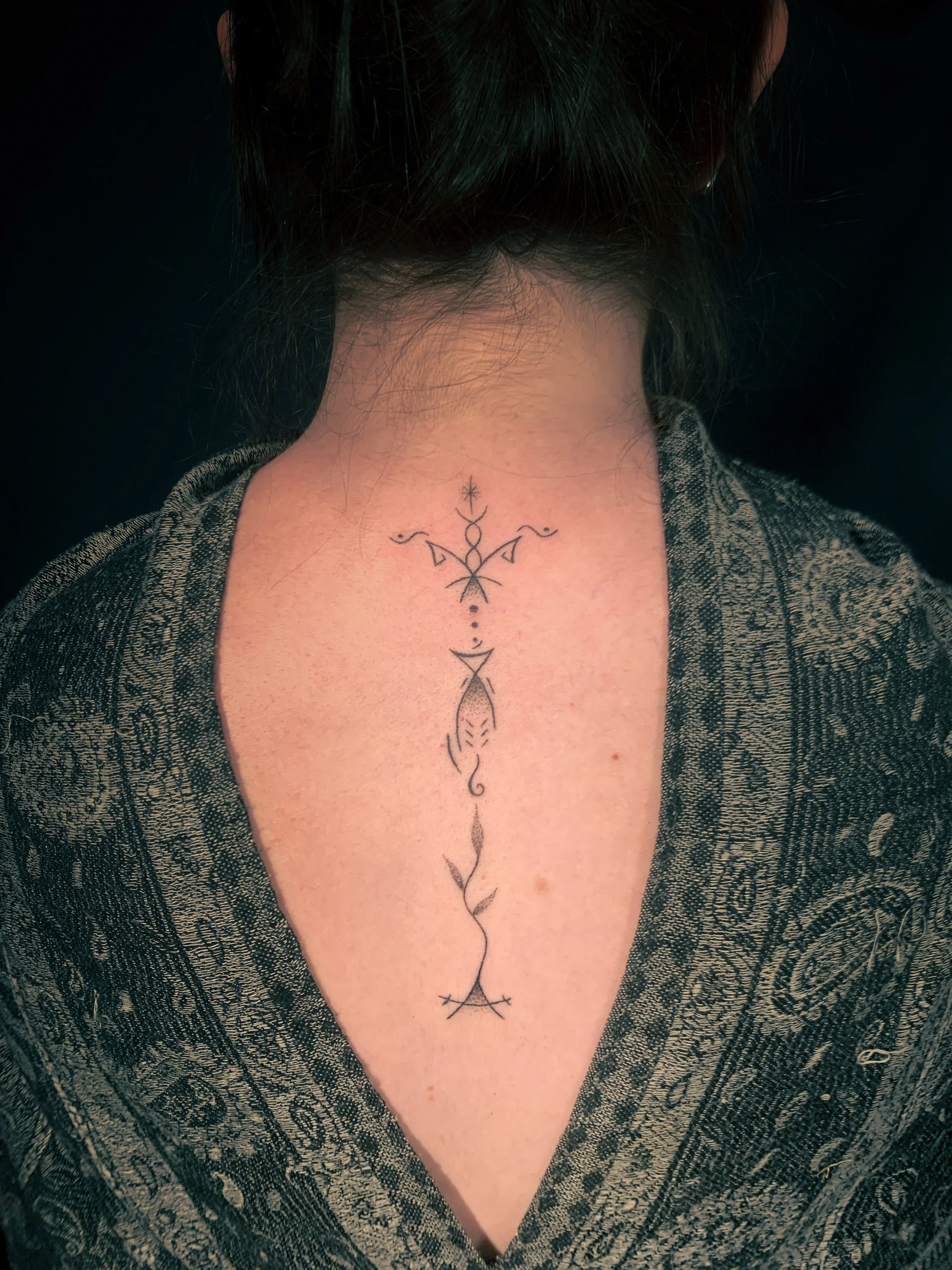 A person's back with a long, thin, decorative tattoo running down the spine, surrounded by dark hair and a patterned garment.