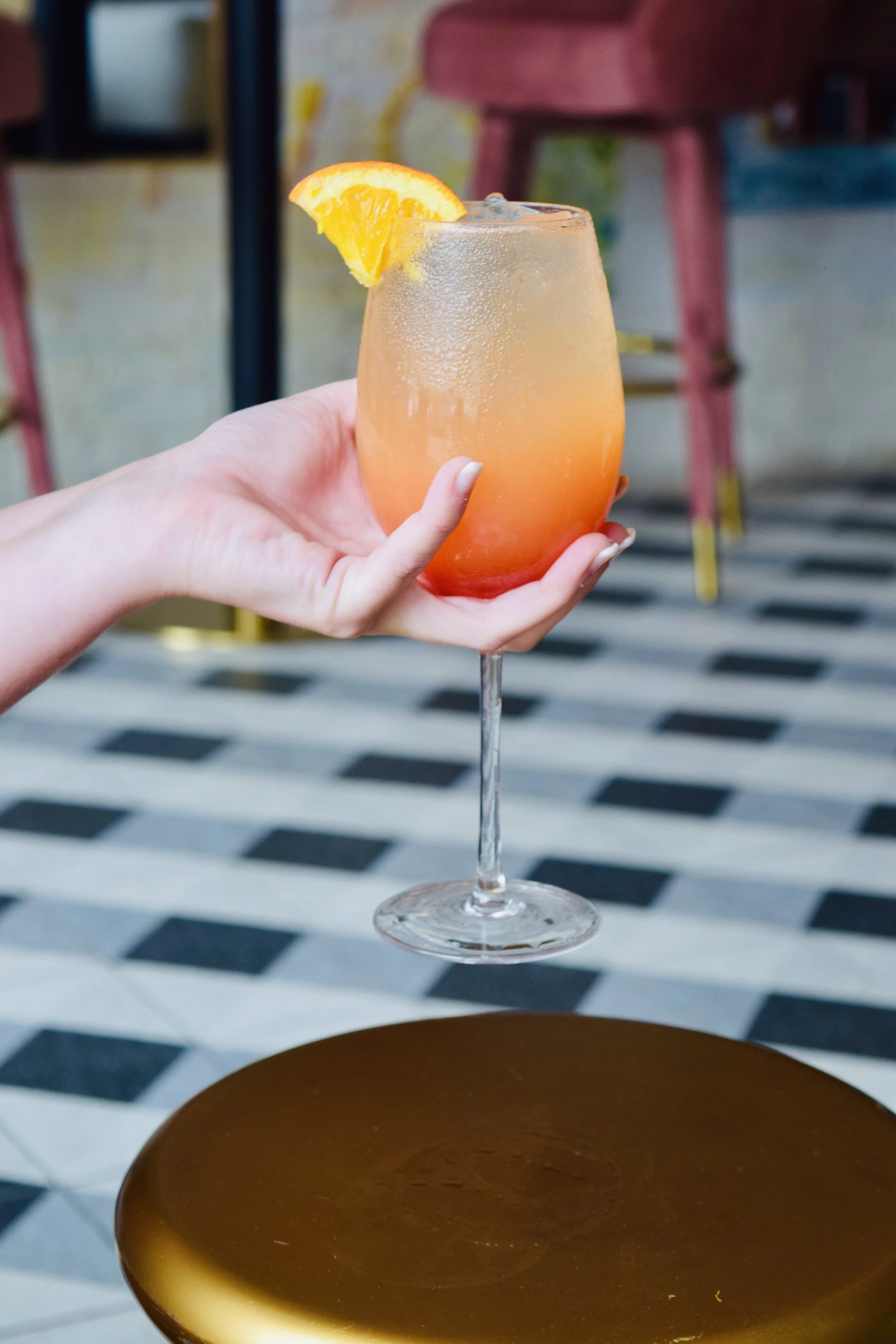 A hand holding a glass of orange-colored cocktail with a slice of orange garnishing the rim, against a background of chairs and a checkered floor.