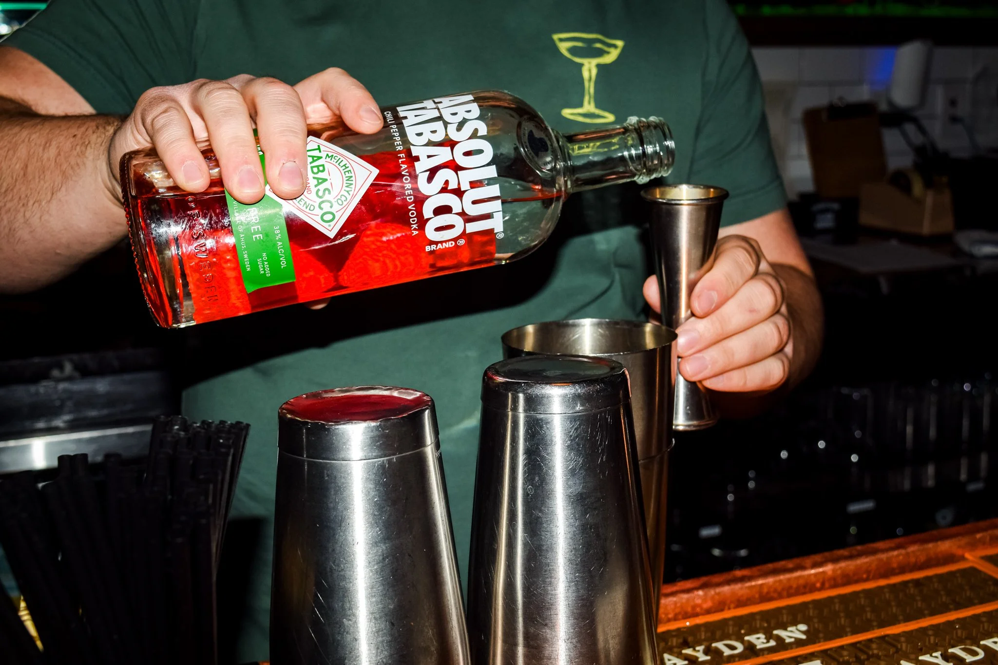 A person pouring red vodka from a bottle labeled 'Absolut Tabasco' into a metal cocktail shaker, with other shakers and bar tools on a bar counter in the background.