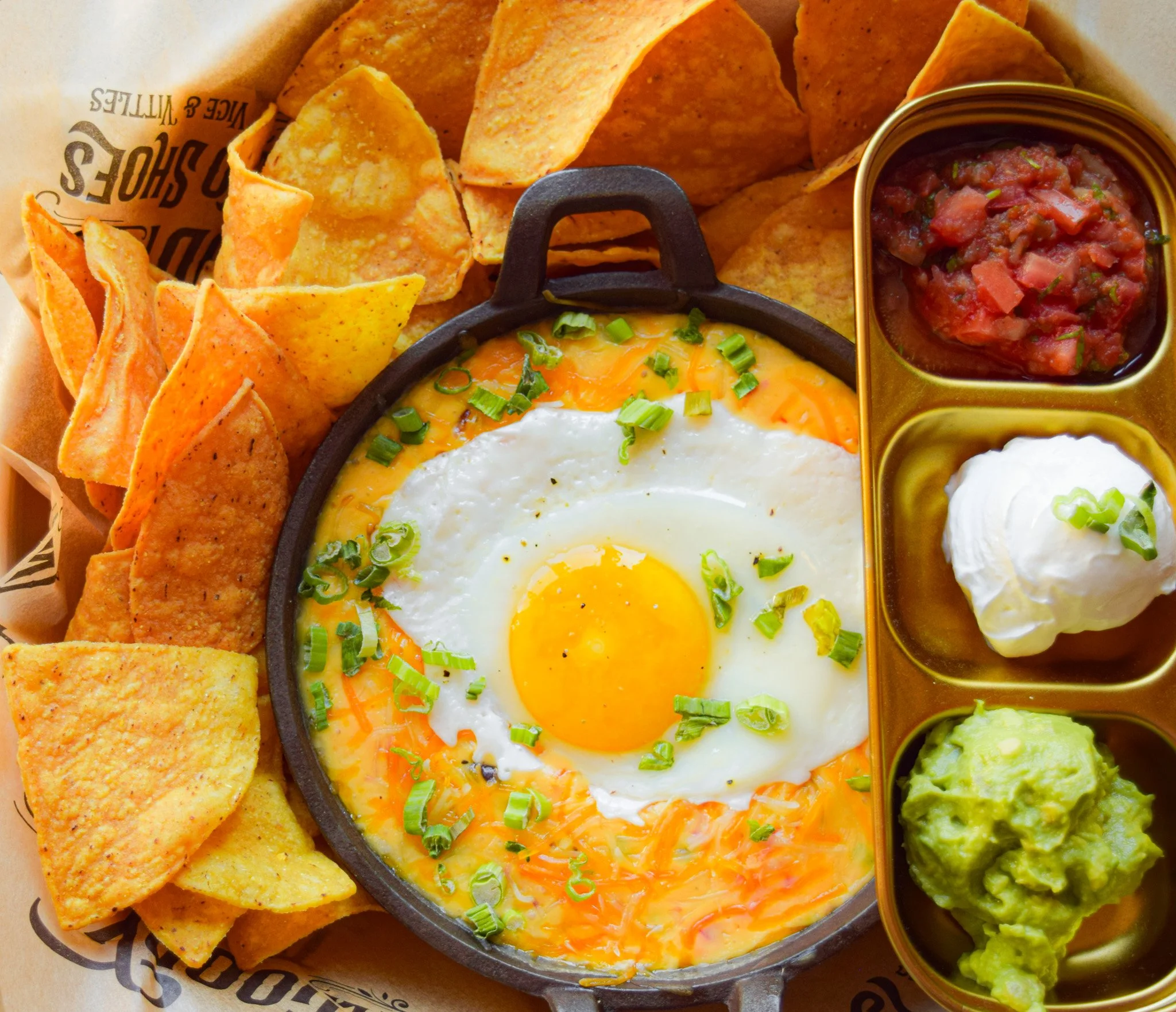 A breakfast dish with a fried egg topped with green onions, served in a skillet with tortilla chips on the side, along with a small metal tray containing salsa, sour cream, and guacamole.