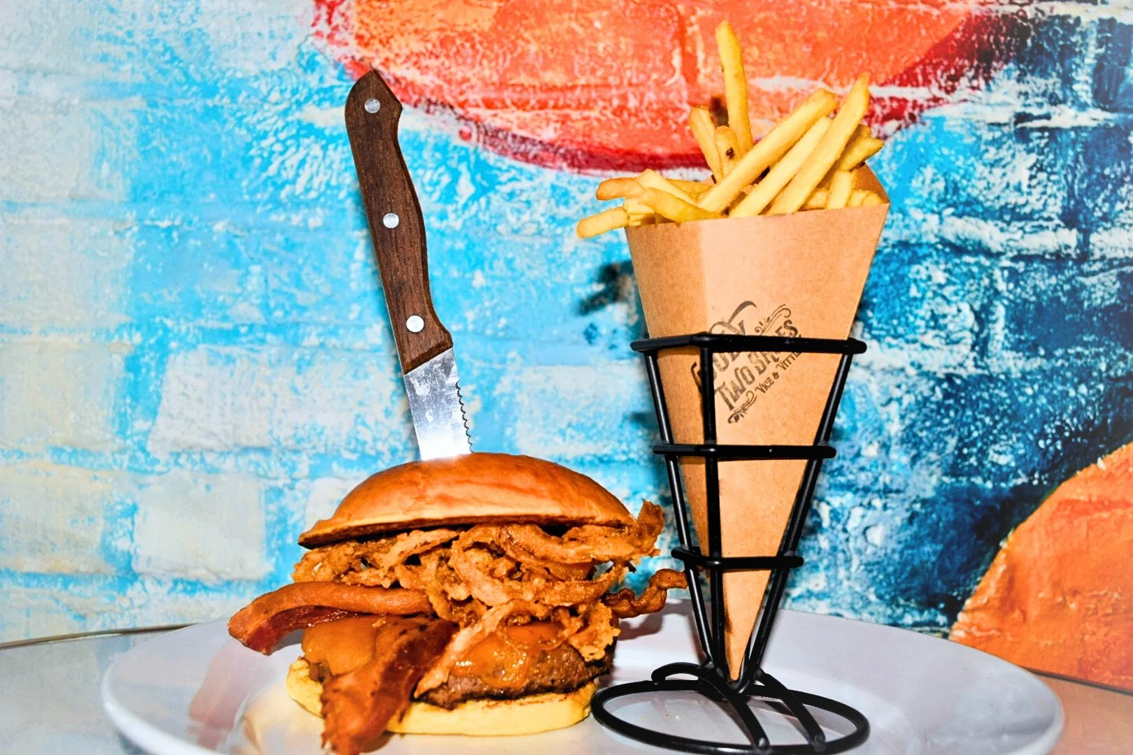 A burger with fried onions and bacon, served on a white plate, with a side of French fries in a cone holder, against a colorful, abstract painted blue and orange background.