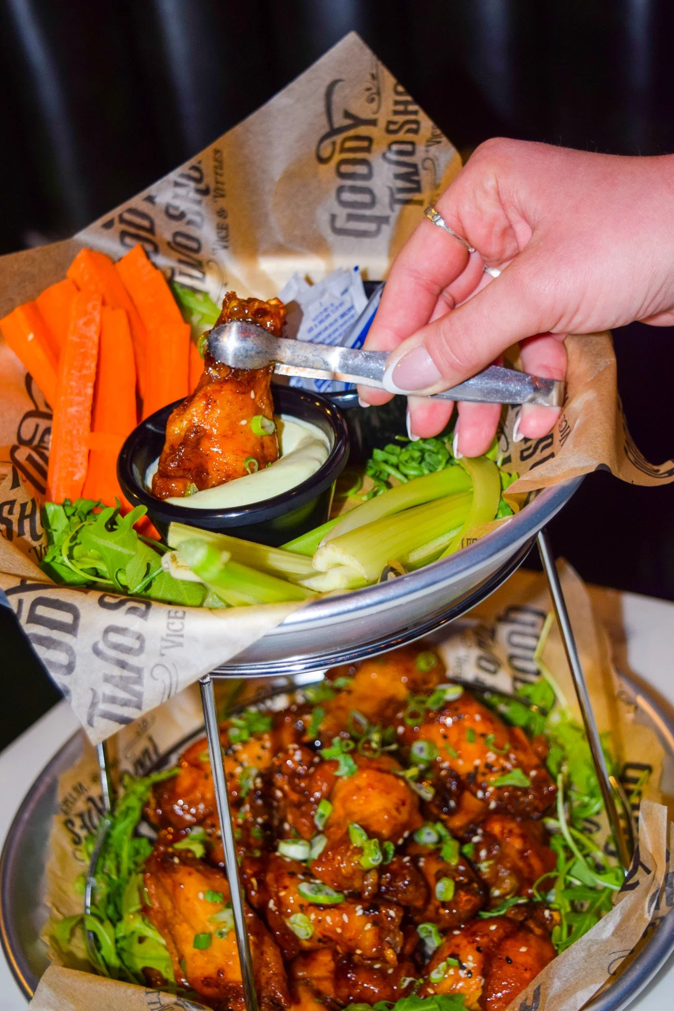 A metal serving tray holding fried chicken drumsticks topped with a dark sauce and chopped green onions, with fresh vegetables including carrots, celery, and lettuce on the side, all served with a small cup of dipping sauce.