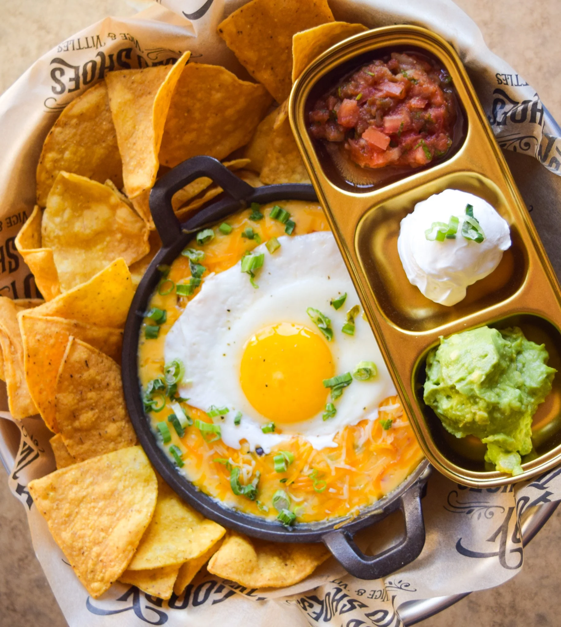 A serving of chips with a good smell. The chips are crispy, and there is a side of salsa, guacamole, and a fried egg with chives. The food is presented on a tray lined with branded paper.