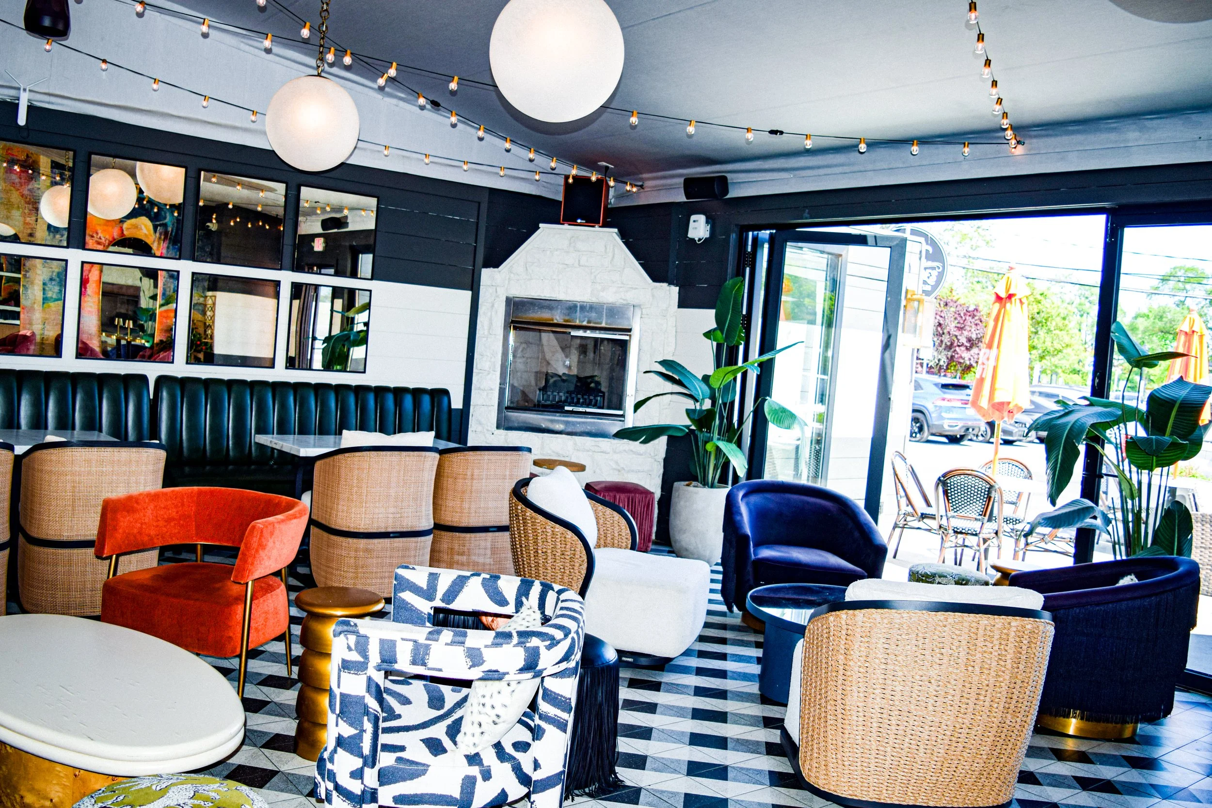 Interior of a stylish cafe with various colorful chairs and sofas, large windows letting in natural light, hanging light bulbs, and a white brick fireplace.