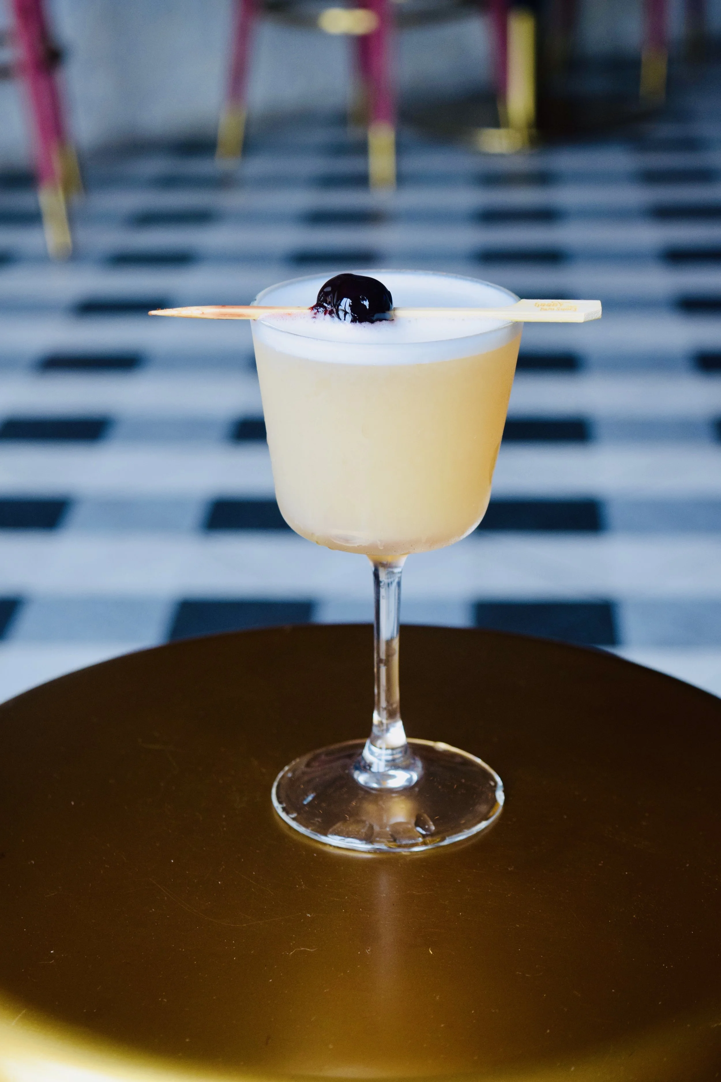 A cocktail glass with a light-colored drink, topped with foam and a dark cherry on a pick, on a dark brown table with striped chairs in the background.