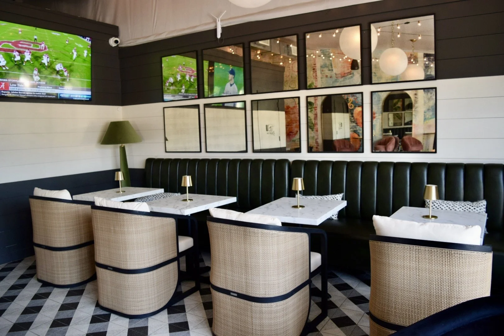 Interior of a modern restaurant with a row of four marble-top tables with beige woven chairs and a black leather banquette. Each table has a small gold lamp, and a green floor lamp is in the corner. The wall is decorated with framed mirrors and glass