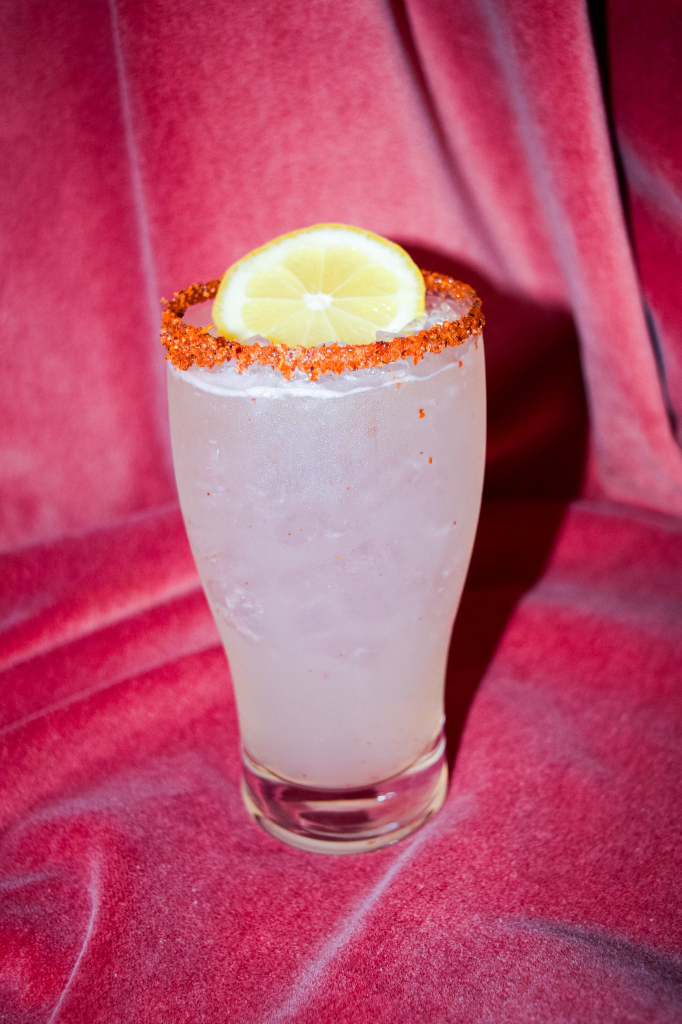 A clear glass filled with a light pink or pale beverage, garnished with a lemon slice on the rim, and an orange chili salt rim. The drink is placed on a red, textured surface.