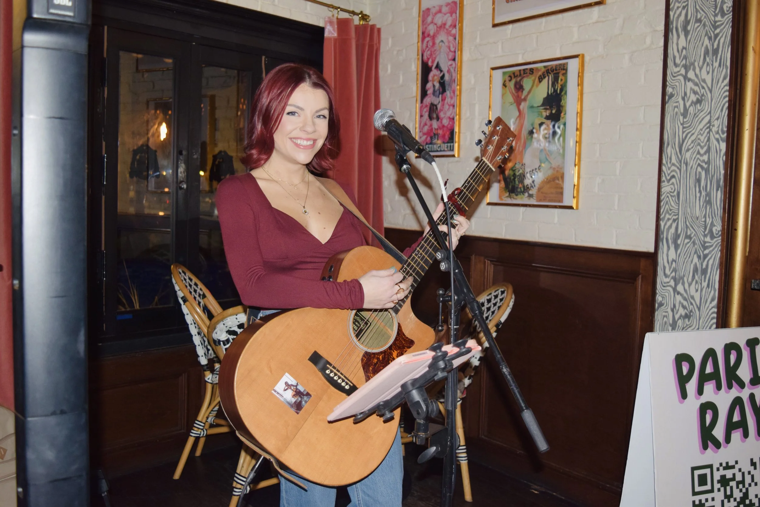 A woman with red hair wearing a maroon top and blue jeans, holding an acoustic guitar and smiling, standing in a cozy indoor setting with framed posters on the wall and a music stand in front of her.