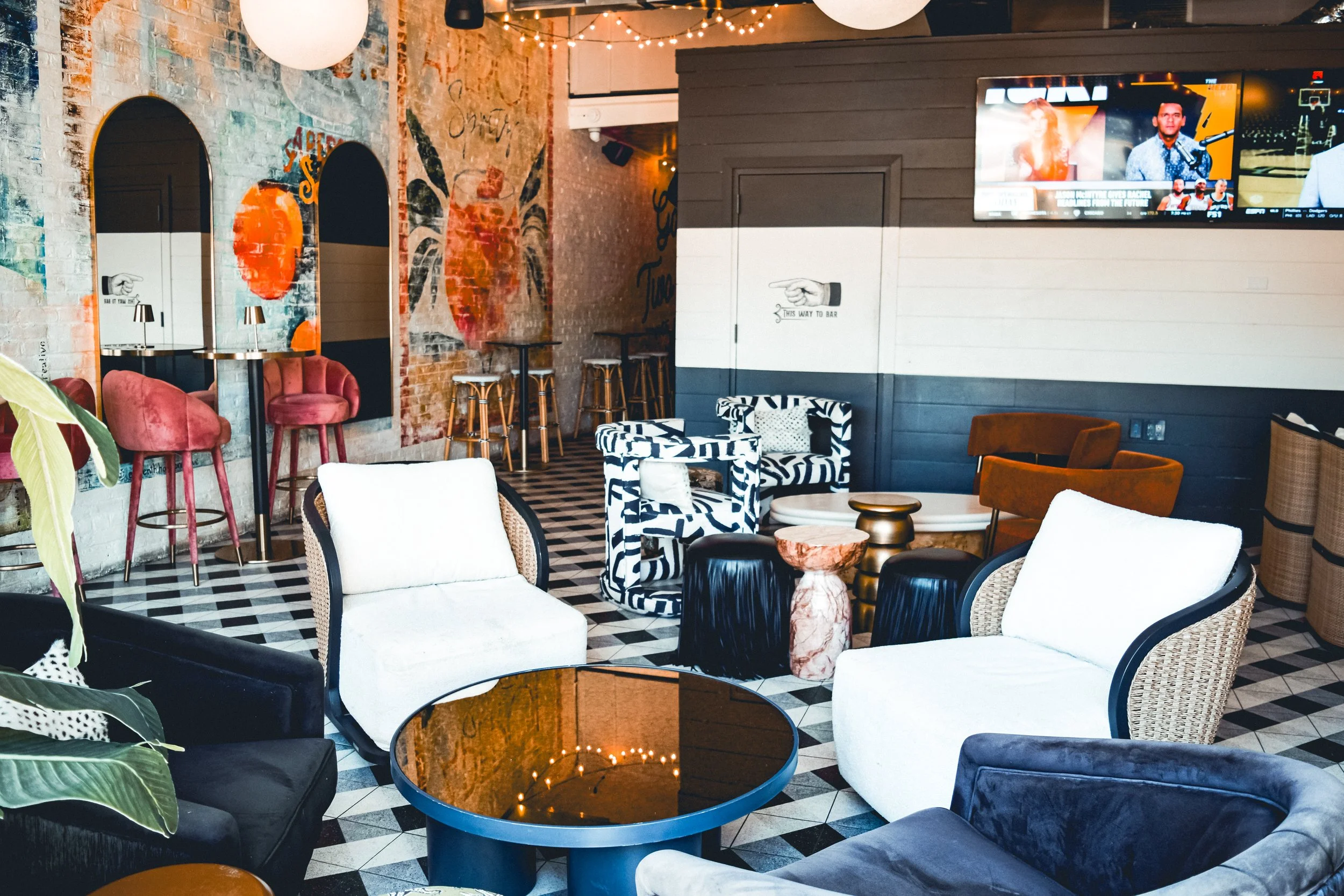 Interior of a modern lounge or bar with a mix of colorful and neutral furniture, including white and black armchairs, pink velvet chairs, and animal print chairs, along with a black and white checkered floor, exposed brick walls with graffiti art, st