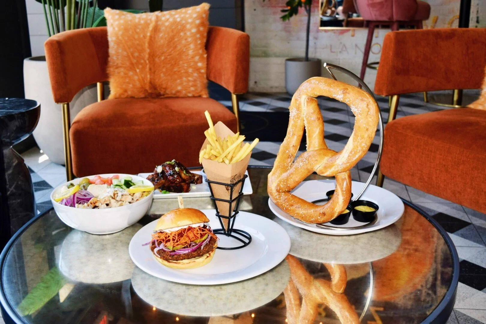 A table with various foods including a salad, fried chicken, French fries, a hamburger sandwich, a large pretzel with mustard, and a small container of dipping sauce, in a cozy restaurant setting with orange chairs.