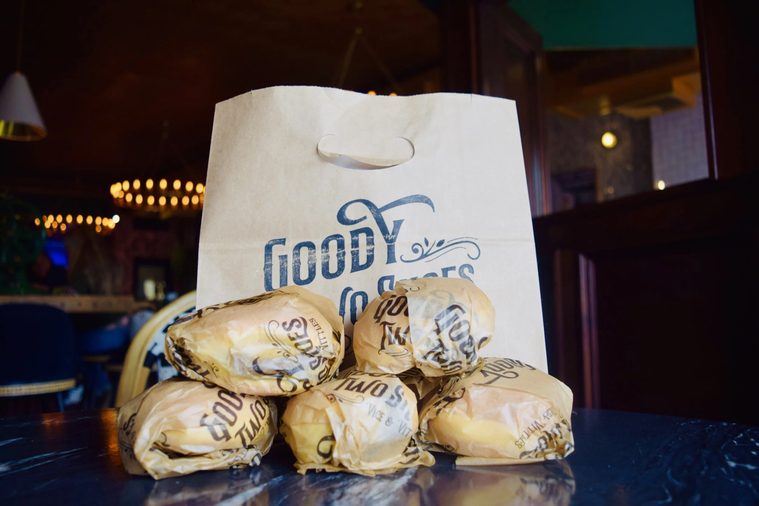 A paper bag with the words "Good Vibes" printed on it, placed on a table with several wrapped sandwiches or burgers in front of it. The background shows a dimly lit restaurant interior with hanging lights and wooden decor.