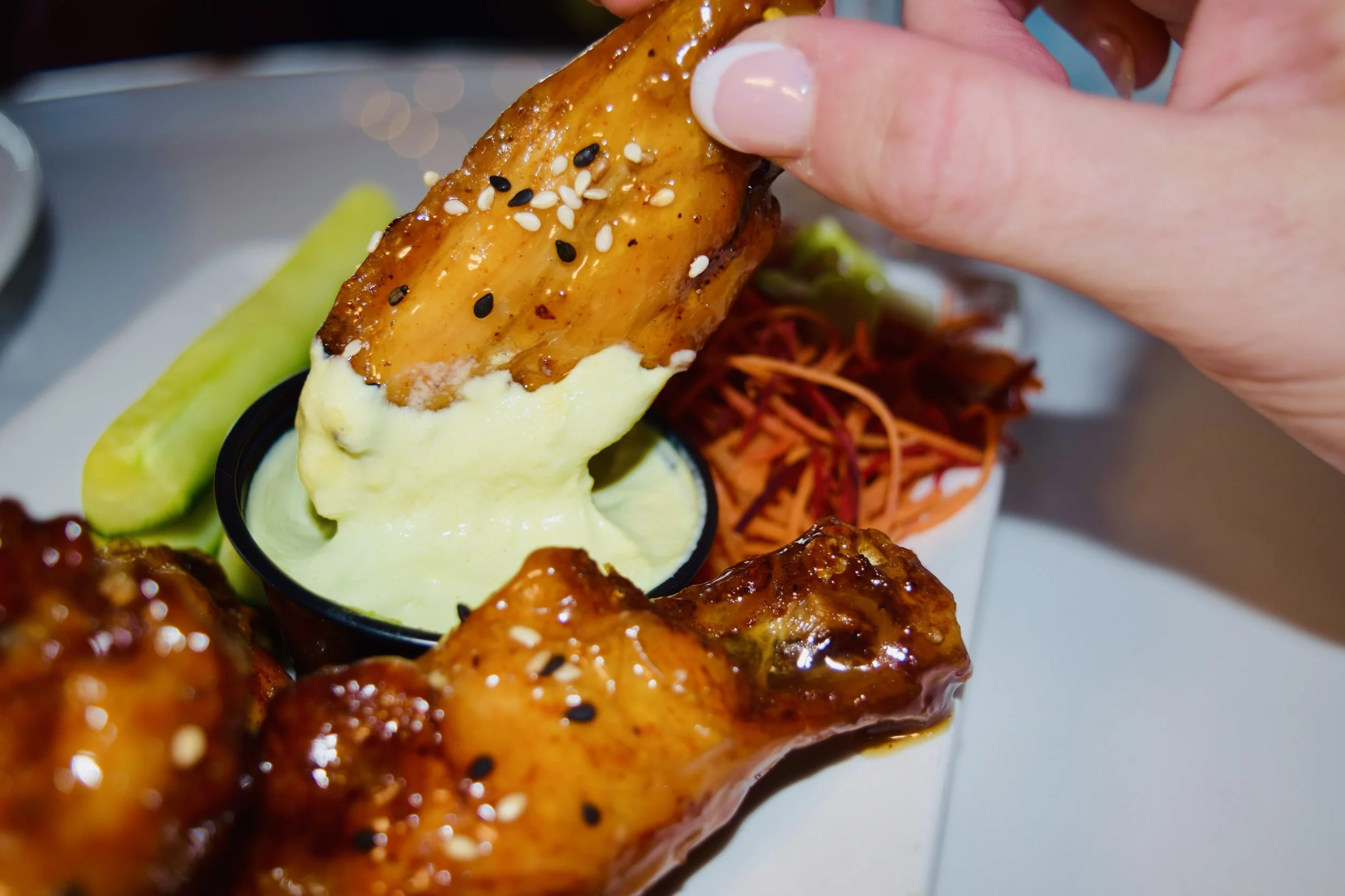 Close-up of a hand holding a piece of glazed chicken wing sprinkled with sesame seeds, with sliced cucumber and shredded carrots in the background.