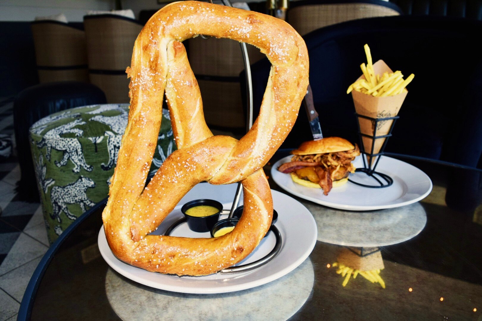 A large salted pretzel on a white plate with small cups of mustard, a burger with fried onions and bacon on a bun, and a cone of French fries on a tray in a restaurant setting.