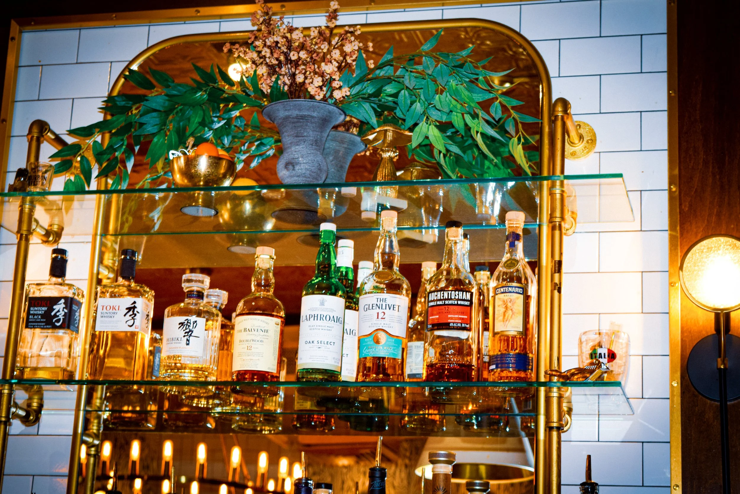 A glass shelf with various bottles of whiskey and other spirits, topped with a decorative floral arrangement in a gray vase and gold containers, set against a background of white tile wall with gold accents.