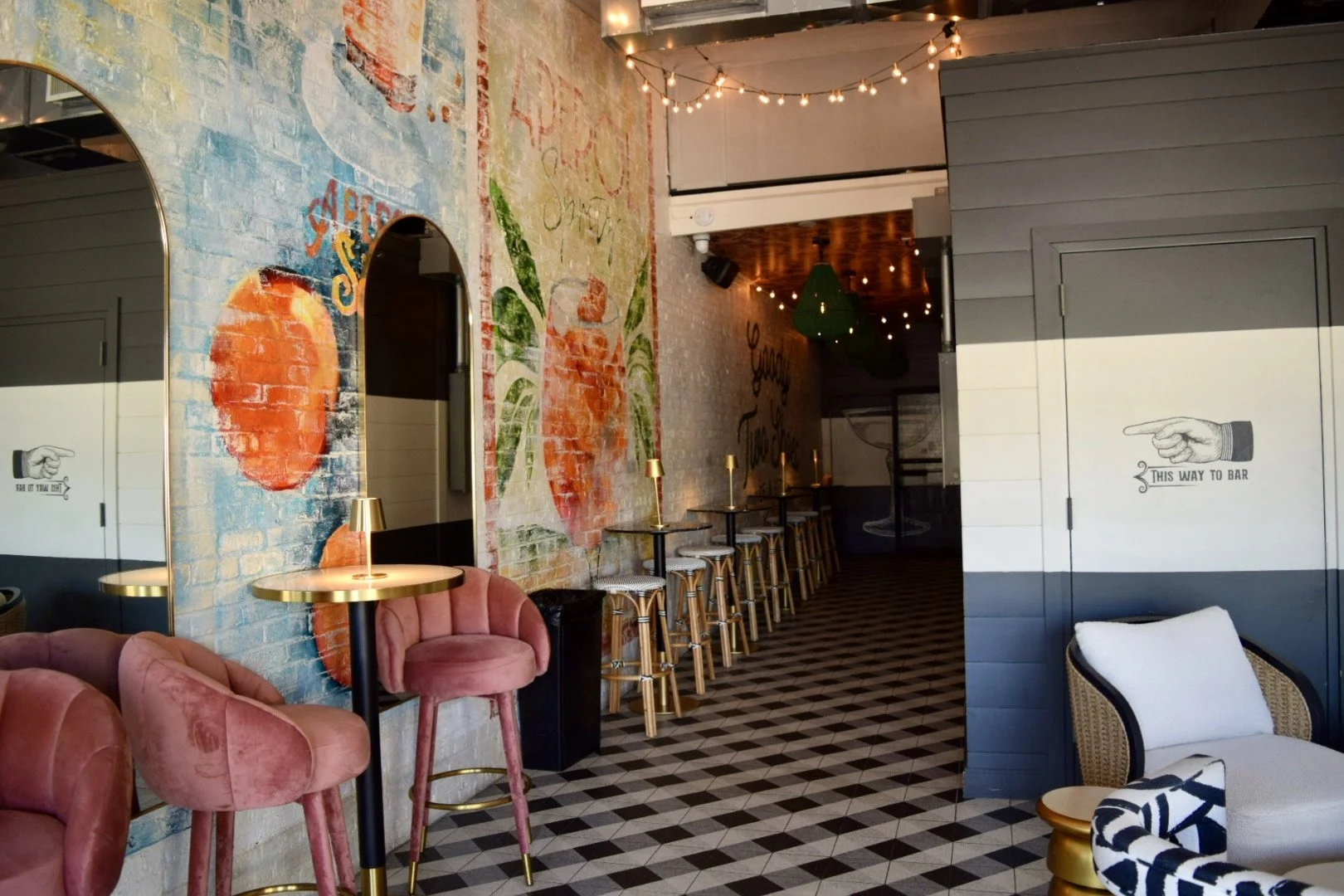 Interior of a modern cafe with pink velvet chairs, a mural with food and drink illustrations, and bar-height tables with barstools along a wall. Decorative string lights hang from the ceiling.