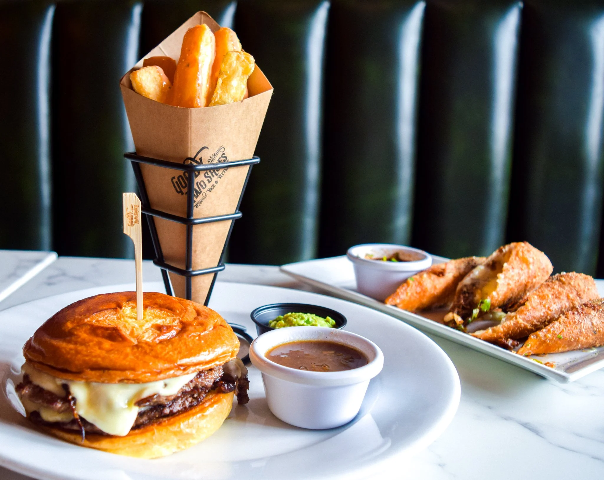 A cheeseburger with grilled onions, melted cheese, and bacon on a bun, served with dipping sauces, alongside fried chicken and other dishes on a white plate.