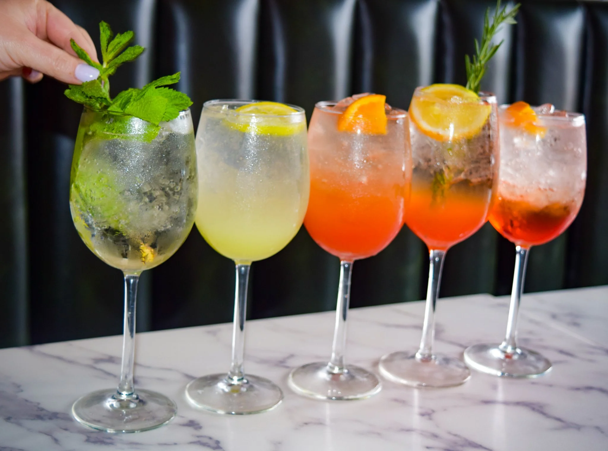 Four colorful cocktails with ice, garnished with citrus slices and herbs, lined up on a white marble surface.