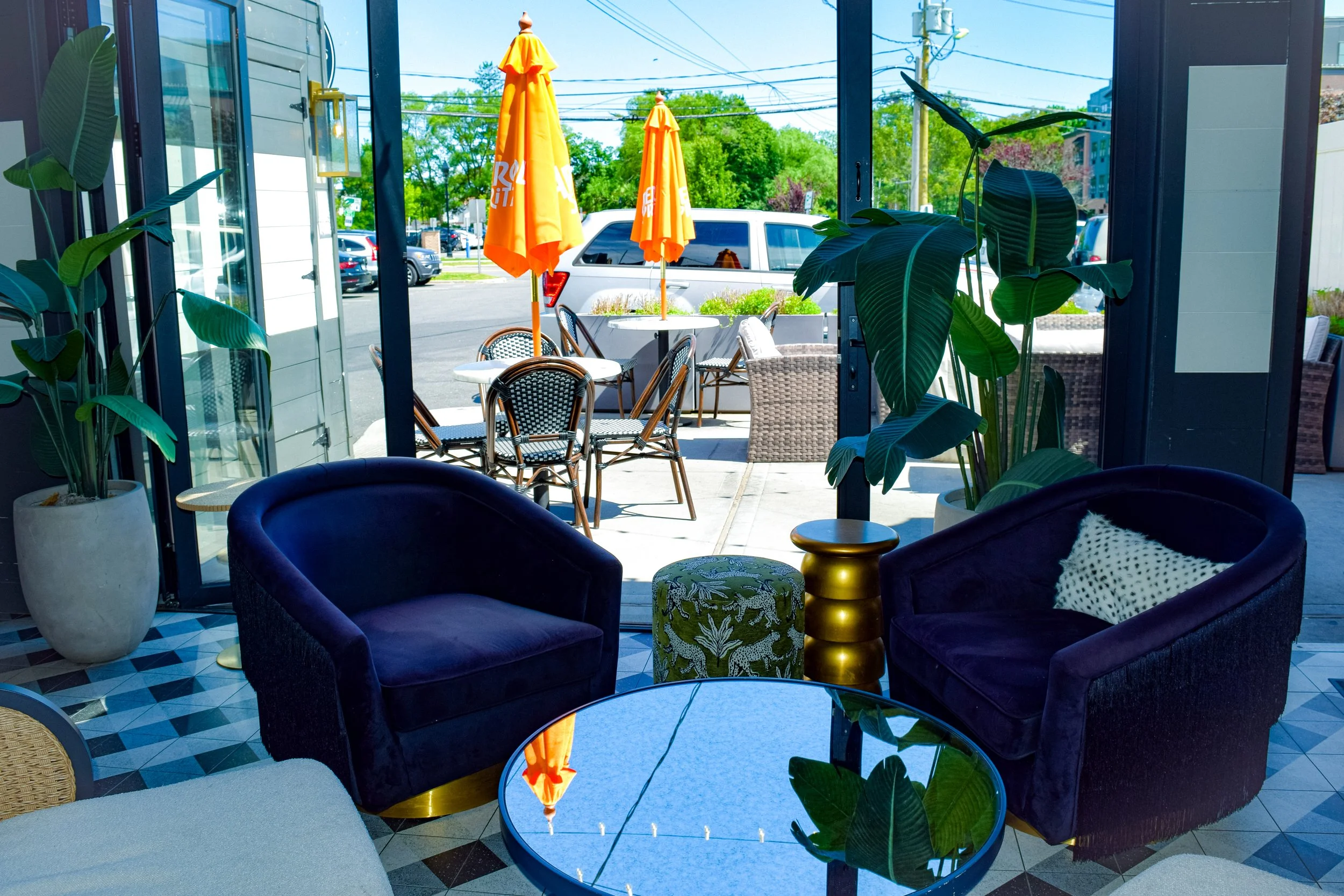 Interior view of a glass-walled cafe with purple velvet chairs, green patterned ottoman, and lots of indoor plants. Outside, there are tables with yellow umbrellas, parked cars, and trees, under a bright blue sky.