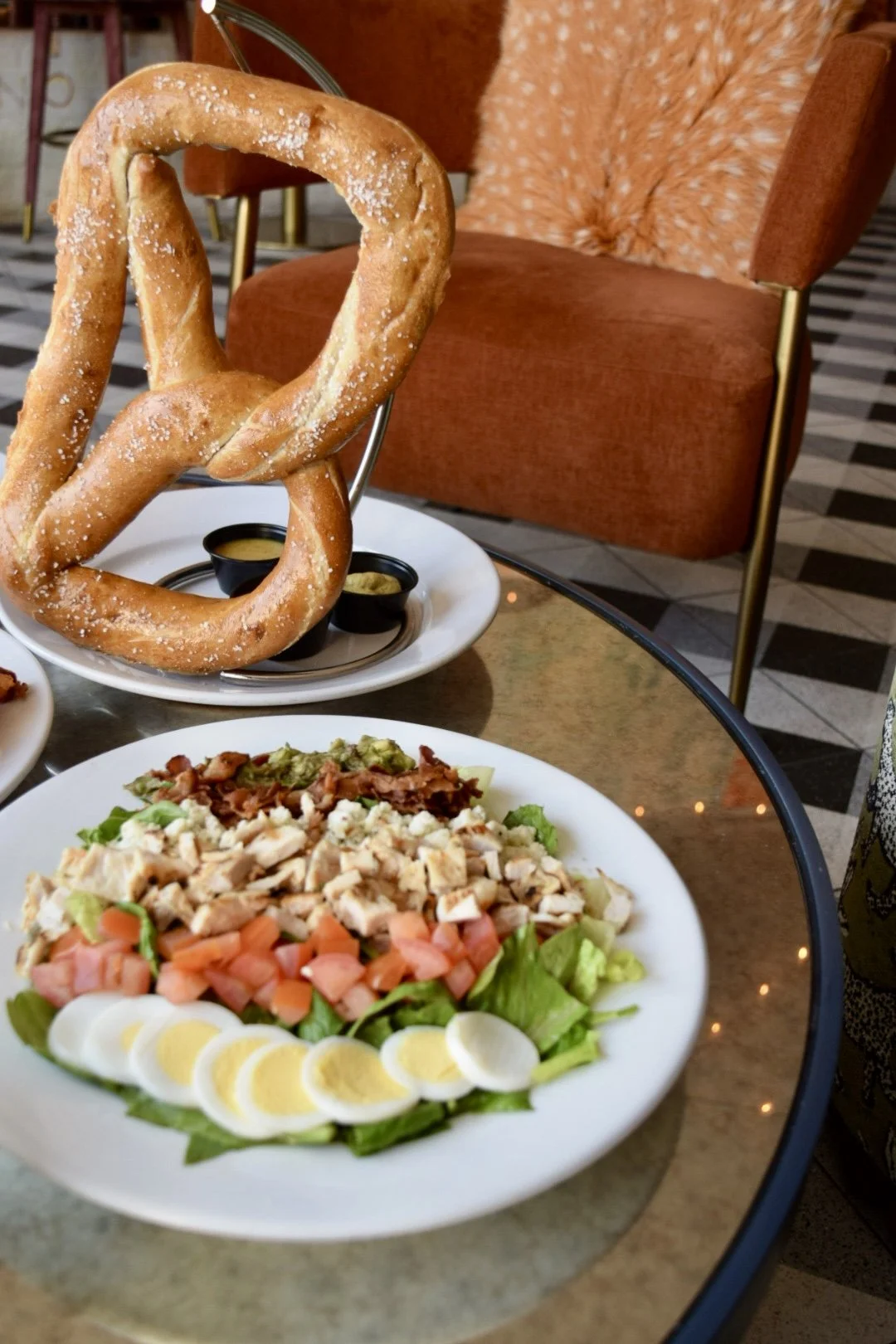 Deep-fried pretzel with salt, served with mustard and cheese sauces, alongside a salad with chopped chicken, tomatoes, boiled eggs, and greens on a white plate, on a glass table in a restaurant setting.