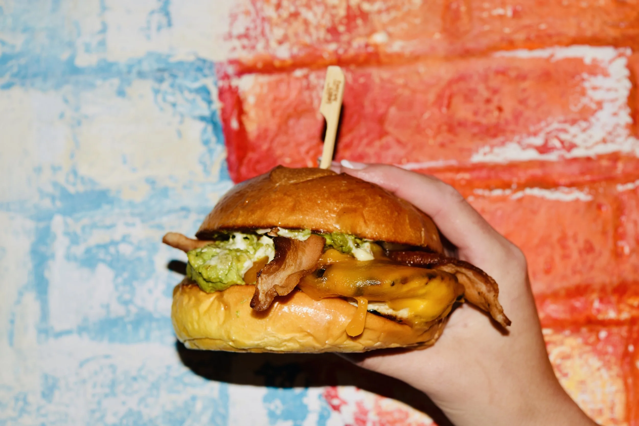 Person holding a gourmet burger with bacon, lettuce, cheese, and a toasted bun, against a colorful abstract background.