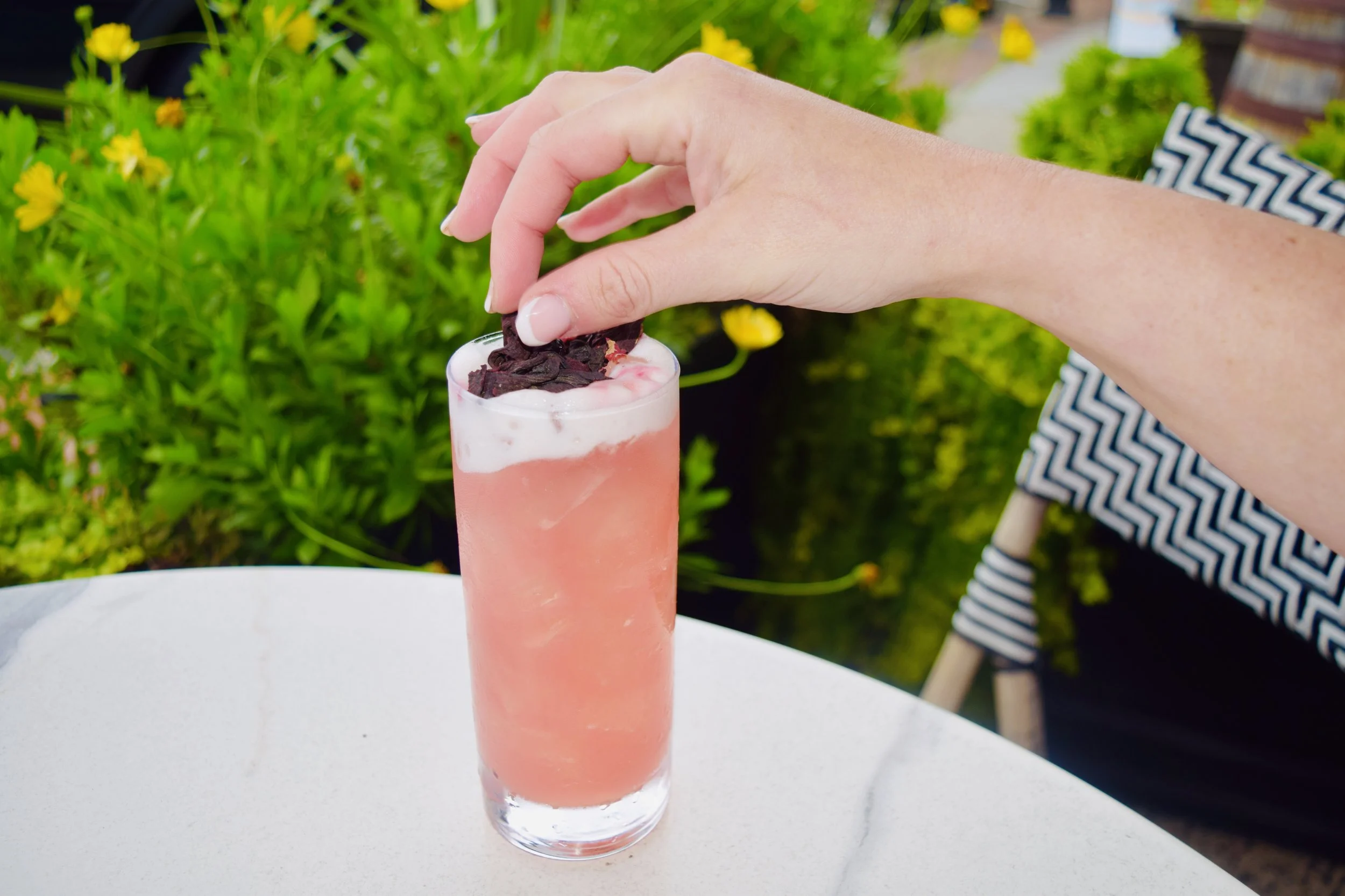 A tall pink cocktail in a glass with whipped cream and dark chocolate shavings on top, set on a white table outdoors with green plants in the background.