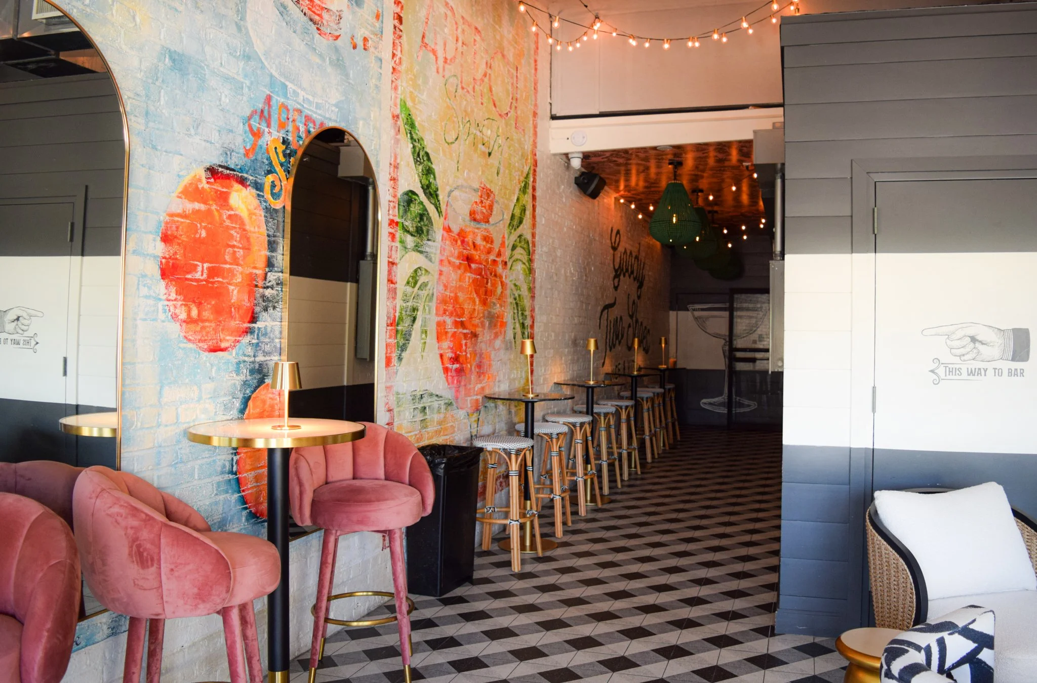 Interior of a stylish bar or café with pink upholstered chairs, gold and black tables, a colorful mural on brick wall, and a row of bar stools along a mural-covered wall. String lights hang from the ceiling, creating a cozy atmosphere.
