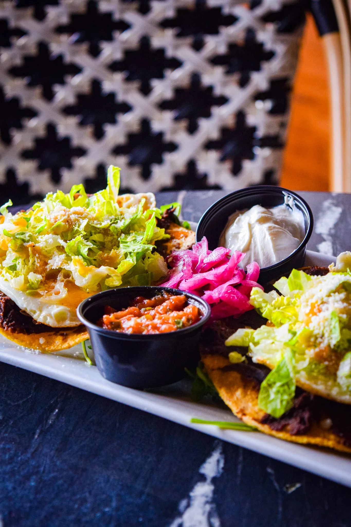 Close-up of a plate of Mexican food, including tacos with lettuce, cheese, and meat, served with small bowls of salsa, sour cream, and pickled red onions on a dark surface.