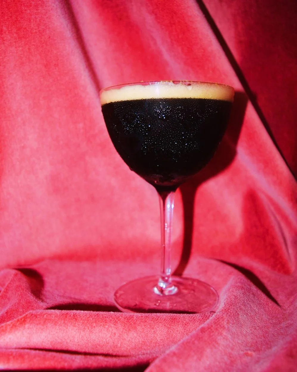 A coupe glass filled with a dark, frothy beverage, set against a pink, velvet background.