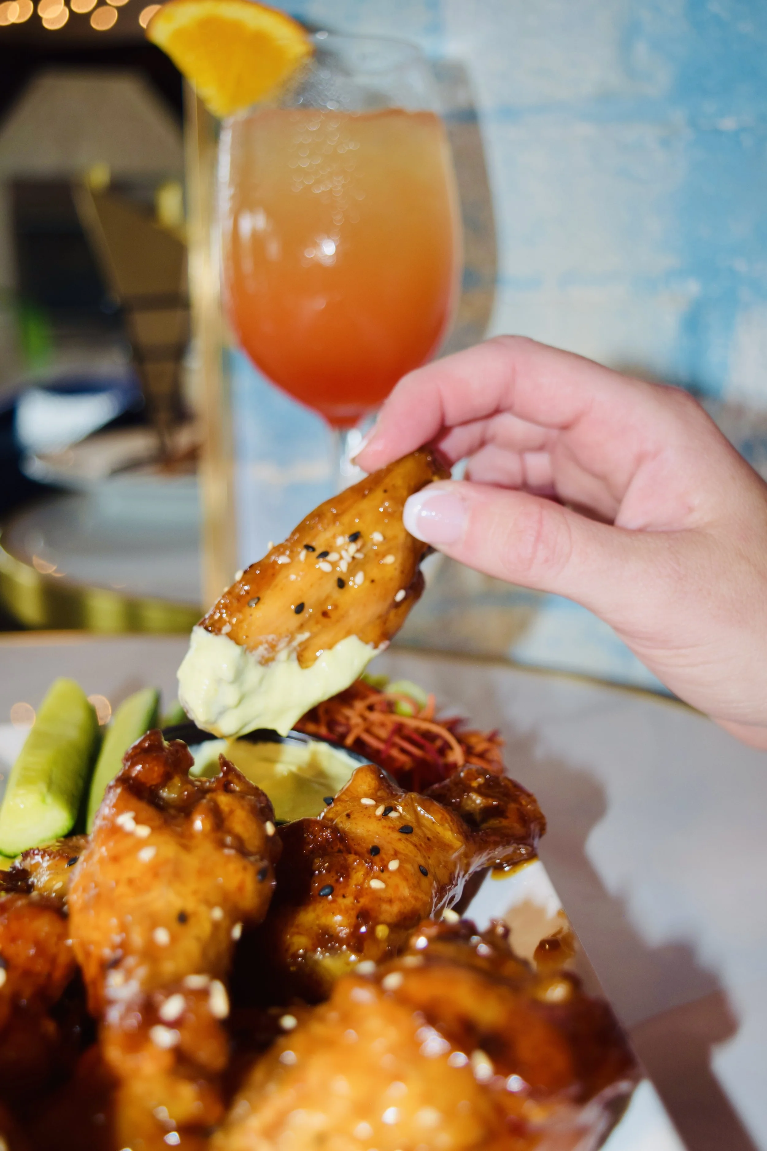 Close-up view of a hand dipping a sesame seed-topped chicken drumstick into a creamy sauce, with a glass of orange-colored drink garnished with an orange slice in the background, and part of a platter with more chicken drumsticks and sliced vegetable