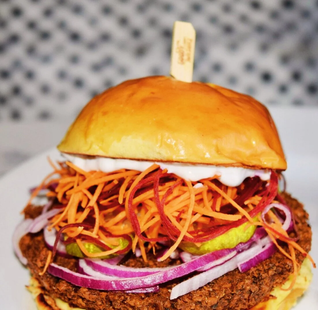Close-up of a burger with a shiny bun, shredded carrots, pickles, sliced onions, and sauce.