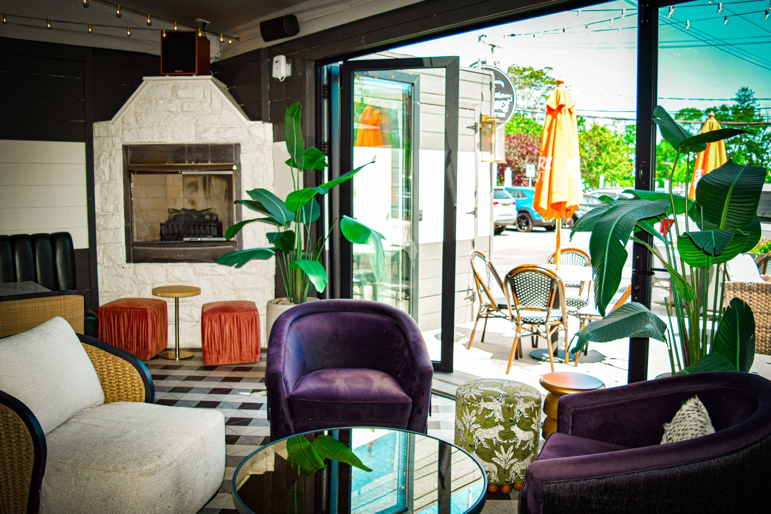 Interior of a cozy cafe with colorful chairs and sofas, large green plants, a fireplace, and a glass door leading outside, with outdoor seating and umbrellas visible.