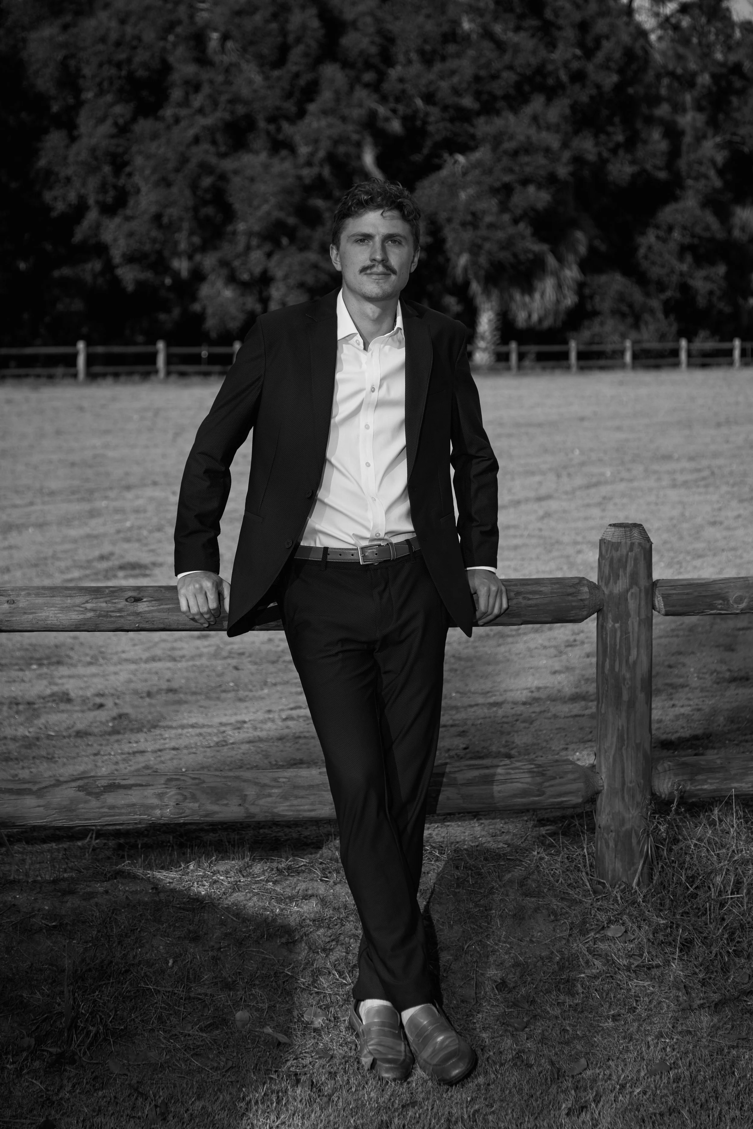 A man dressed in a suit standing outdoors in front of a wooden fence, with trees in the background.