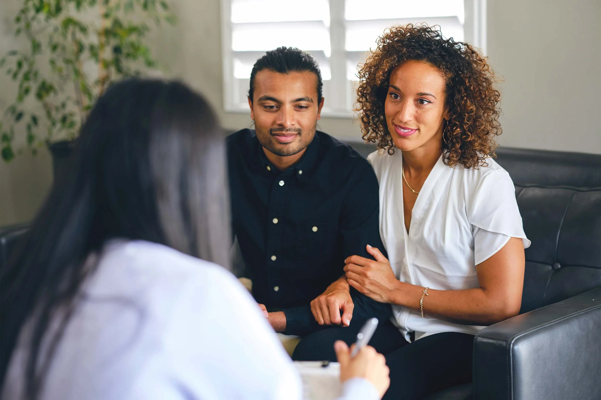 An interracial couple sitting on a black leather couch, talking with a woman who appears to be a therapist or counselor. The woman has curly hair and is wearing a white blouse. The man has long dark hair and is wearing a dark shirt. The counselor has straight dark hair and is taking notes.
