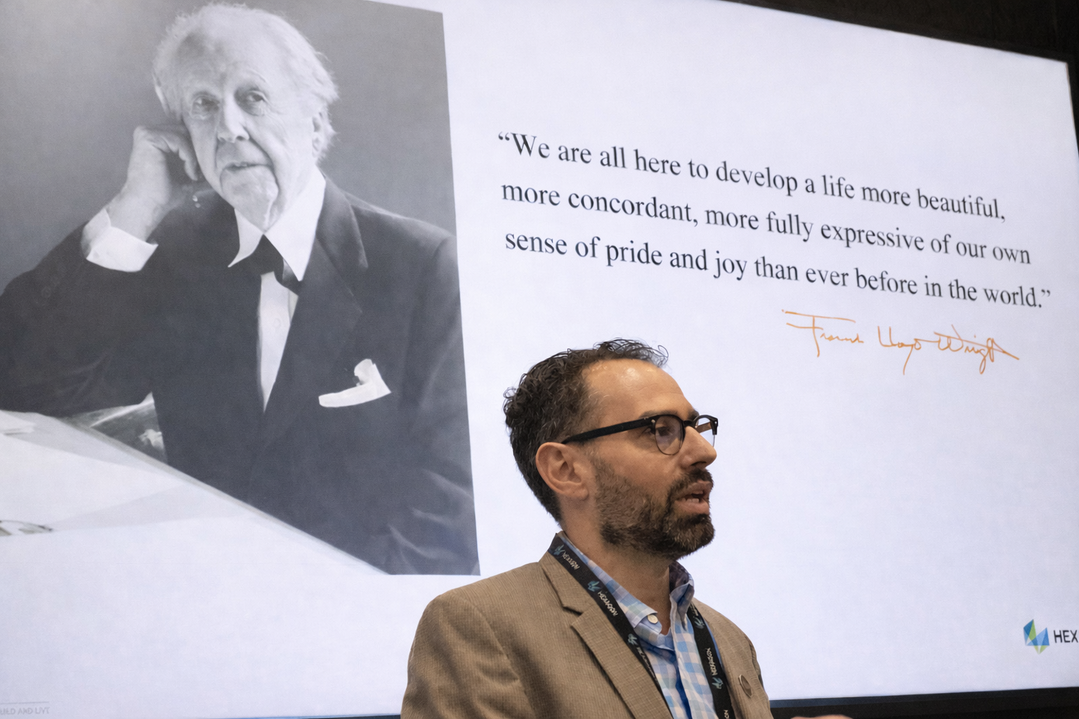 A man with glasses and a beard standing in front of a large screen displaying a black-and-white photo of an elderly man, along with a quote about developing a more beautiful life, attributed to Frank Lloyd Wright.