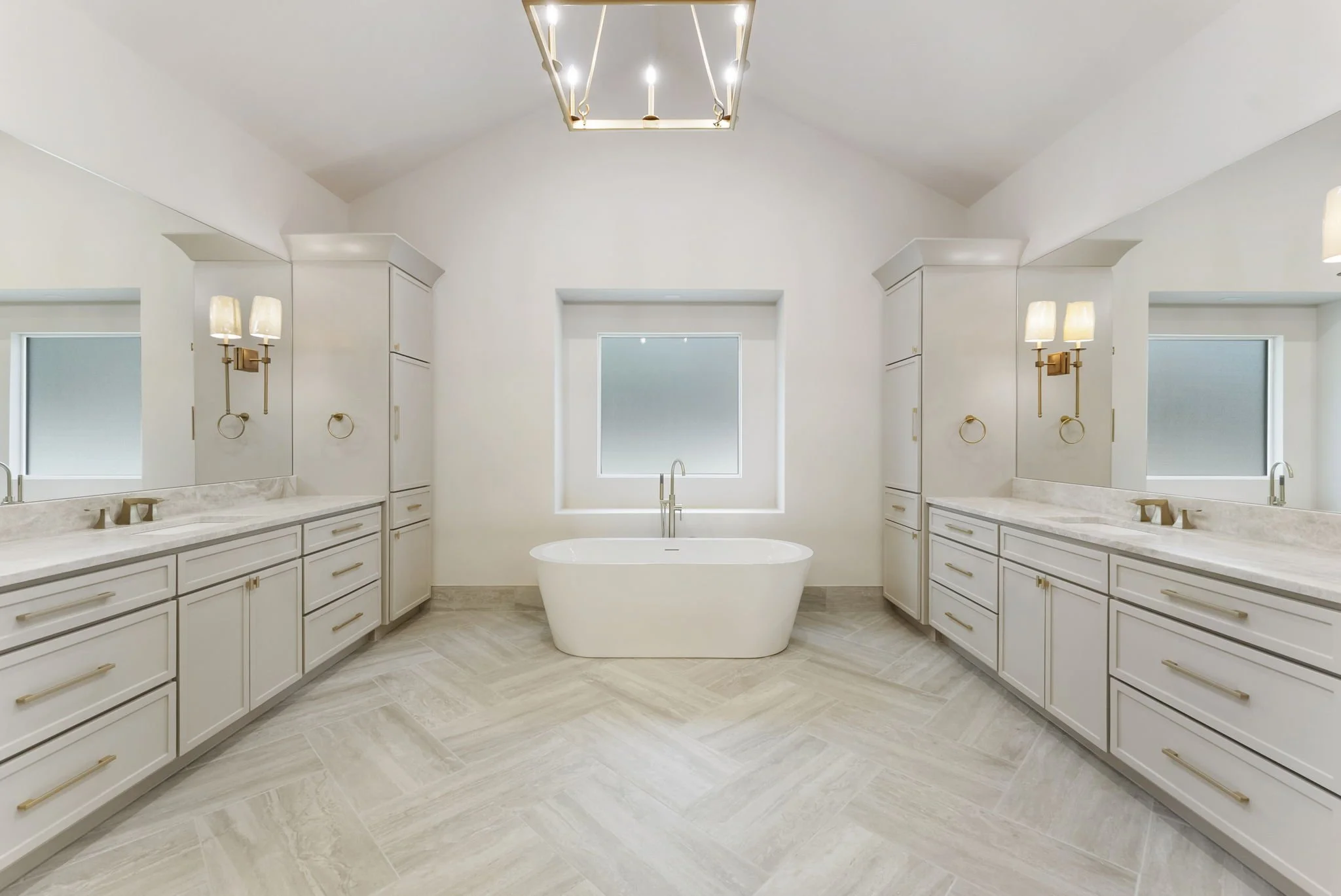 A spacious, modern bathroom with a central bathtub beneath a window, white cabinetry, marble countertops, large mirrors, and elegant wall-mounted lights.
