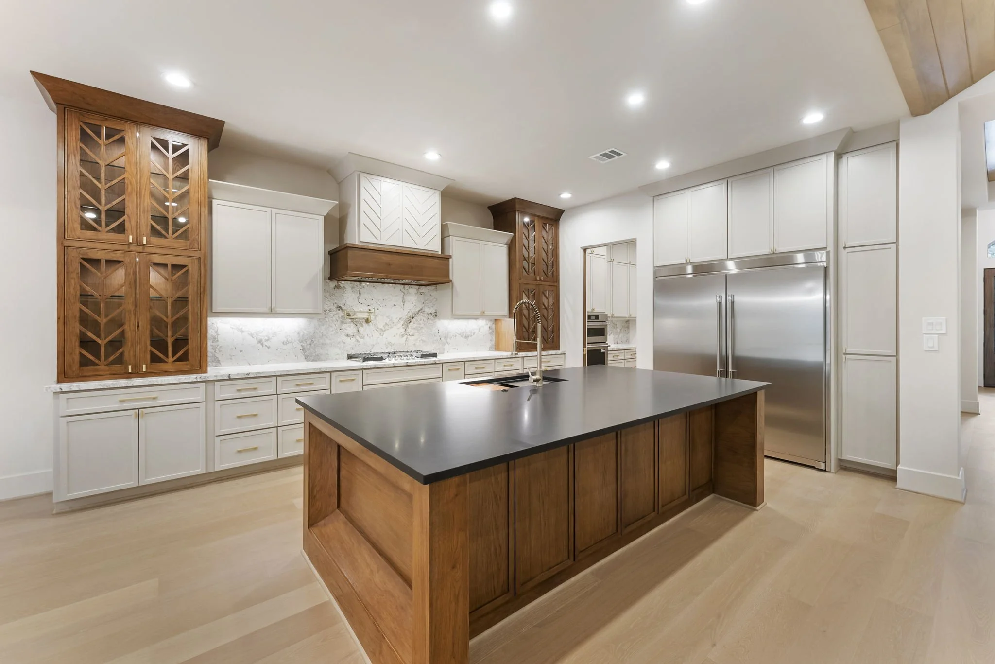 Modern kitchen with white cabinets, a large black countertop island with wooden base, stainless steel refrigerator, and decorative wooden and white cabinetry accents, illuminated by recessed ceiling lights.