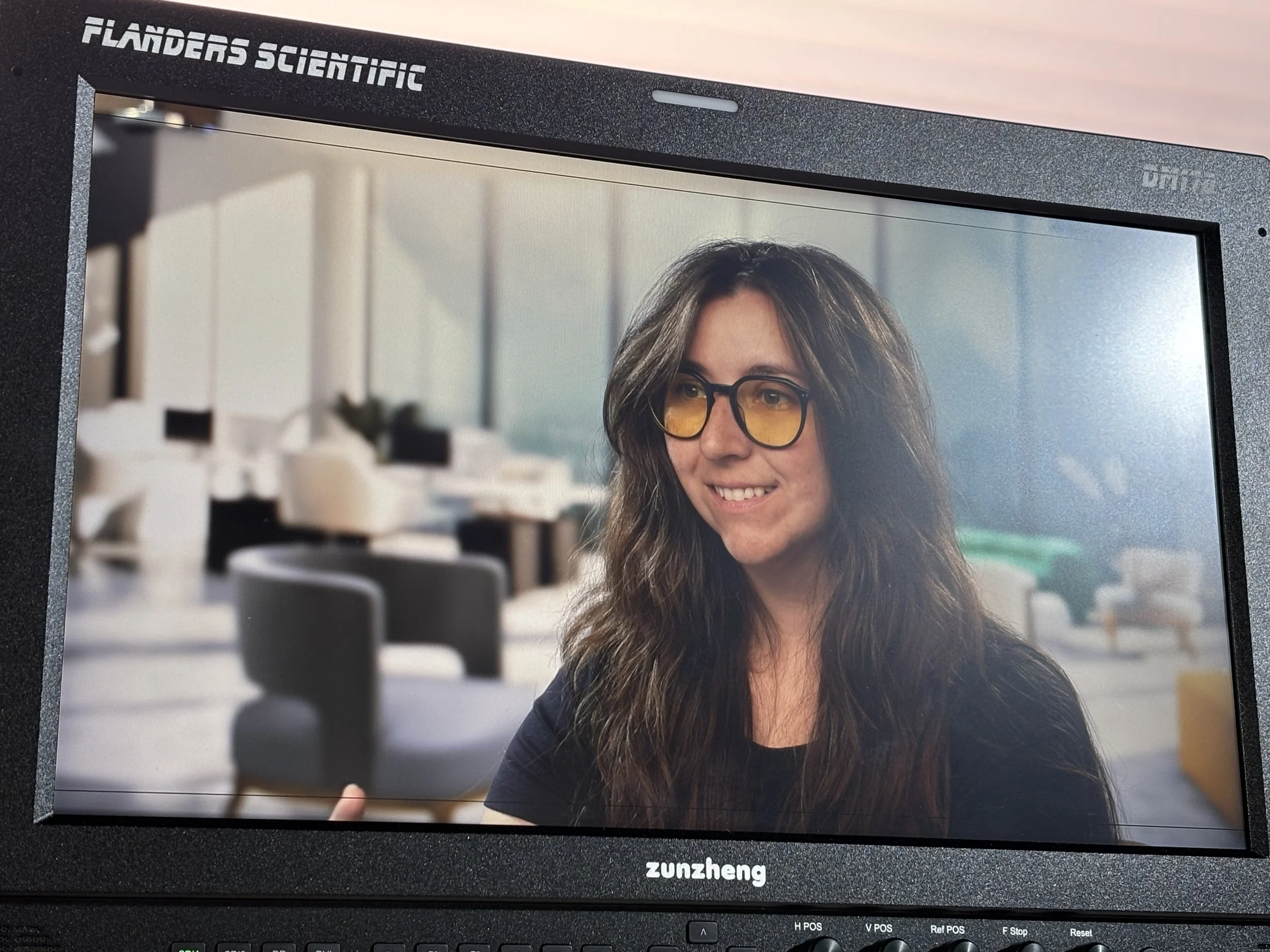 A woman with long wavy hair and yellow-tinted glasses smiling and holding a smartphone. Behind her is a blurred modern office setting with white couches and large windows.
