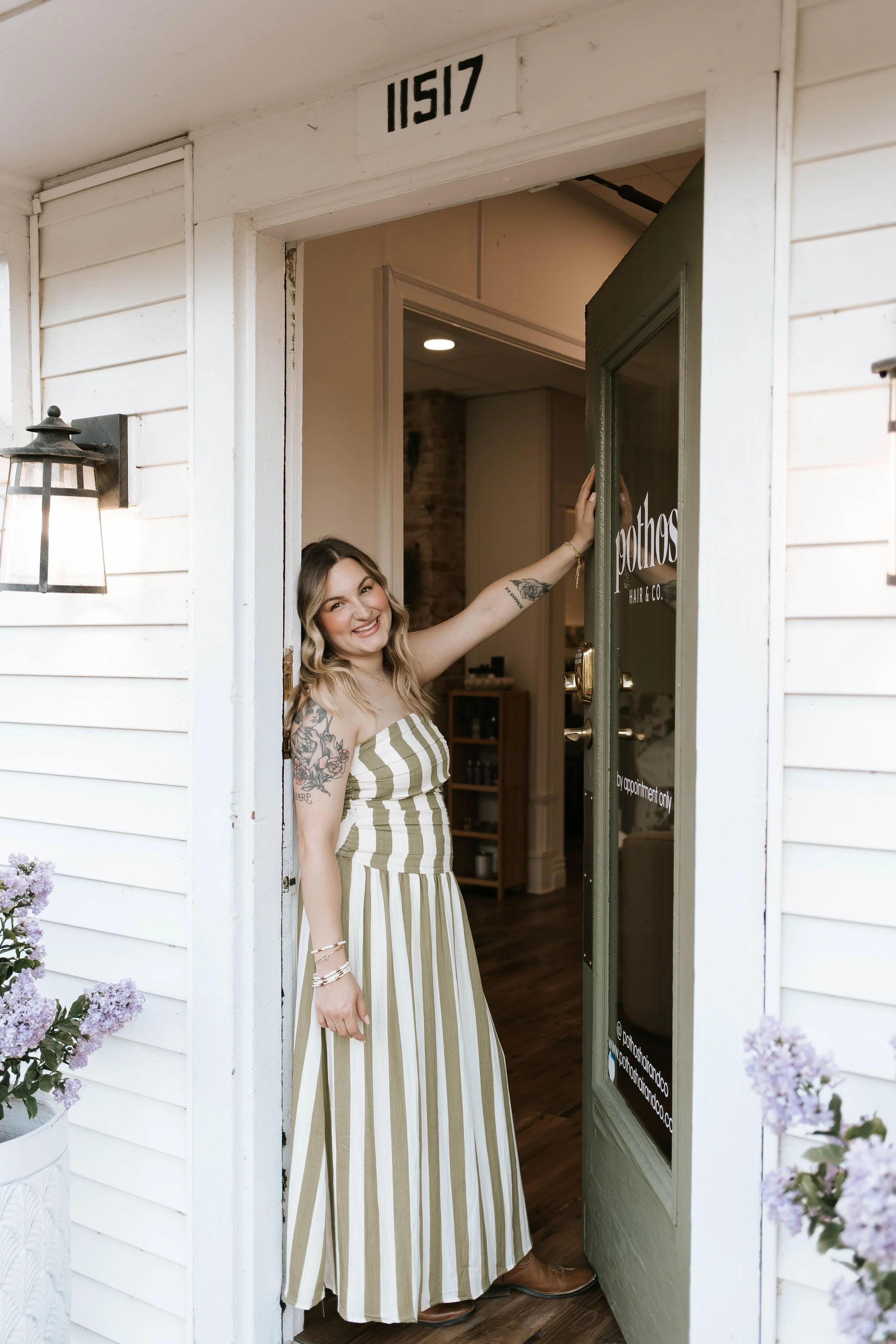 A smiling woman with blonde hair, tattoos on her arm, wearing gold earrings, a white headband, a brown suede vest over a white dress, standing in a greenhouse with green plants and wooden tables.
