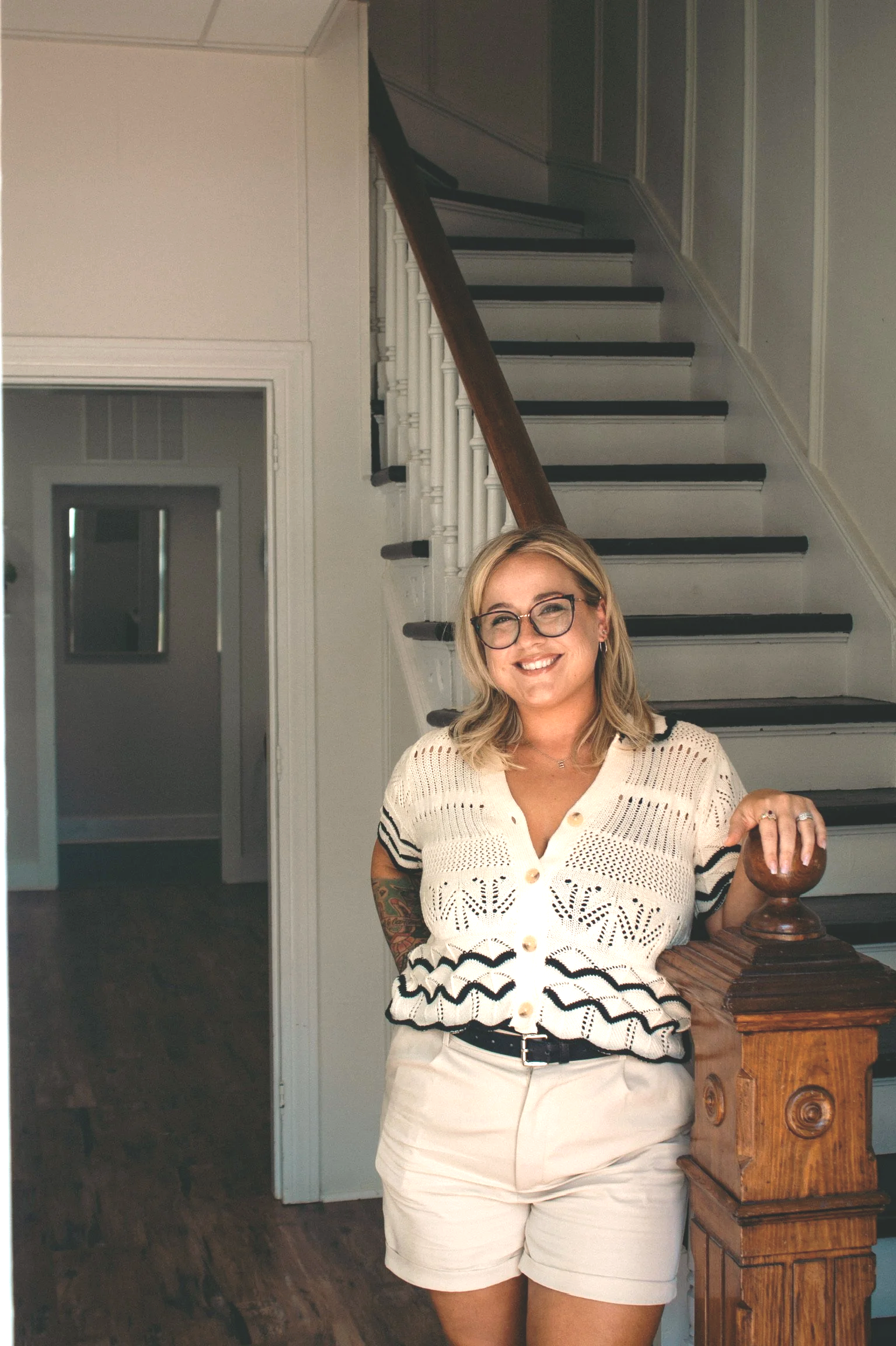 A woman with blonde hair, glasses, and tattooed arm smiling outdoors against a background of green plants and colorful flowers, wearing a white top.