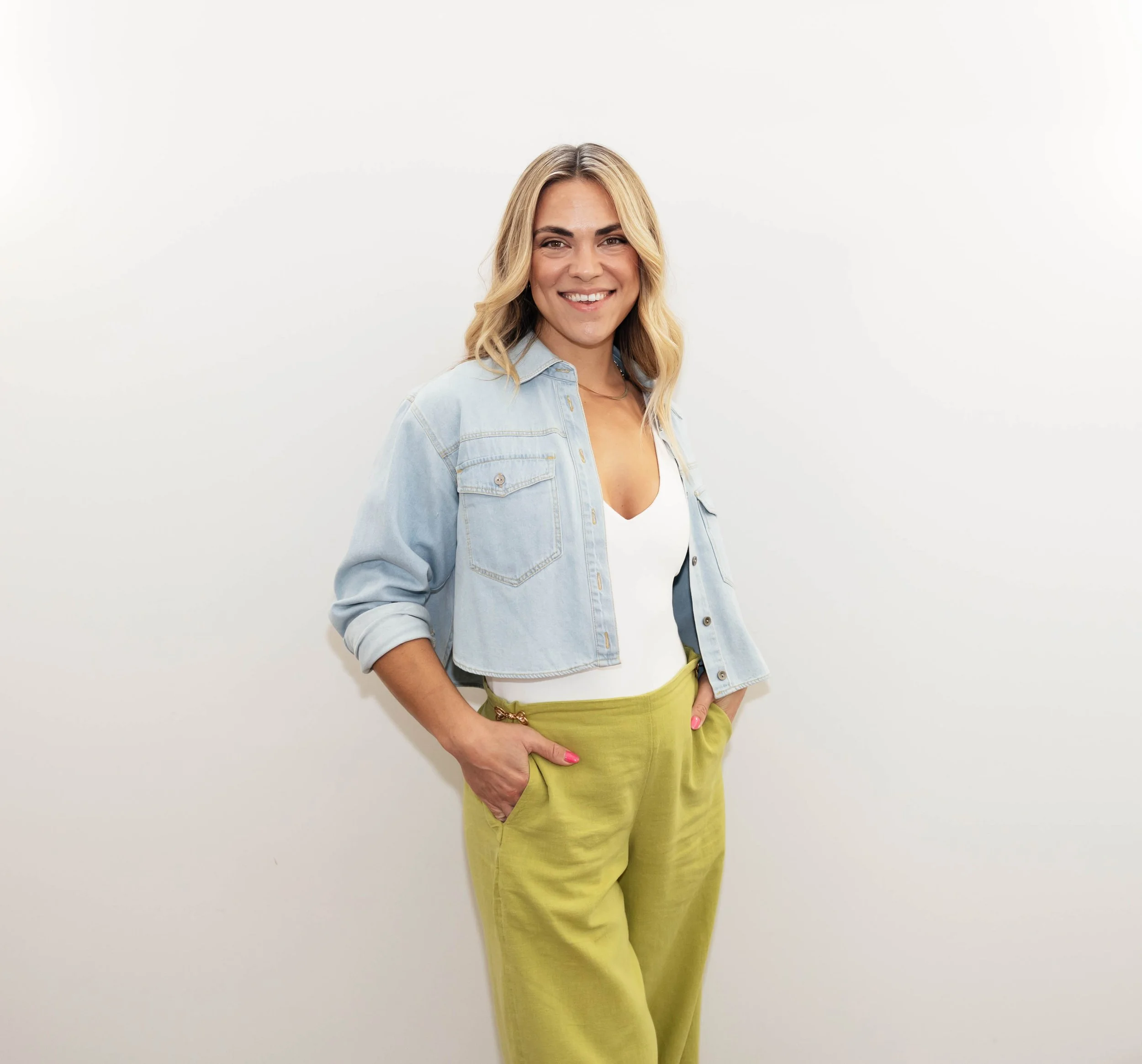 A woman with blonde hair wearing an orange blouse and light blue jeans, standing barefoot against a beige background, smiling and posing with one hand in her pocket.